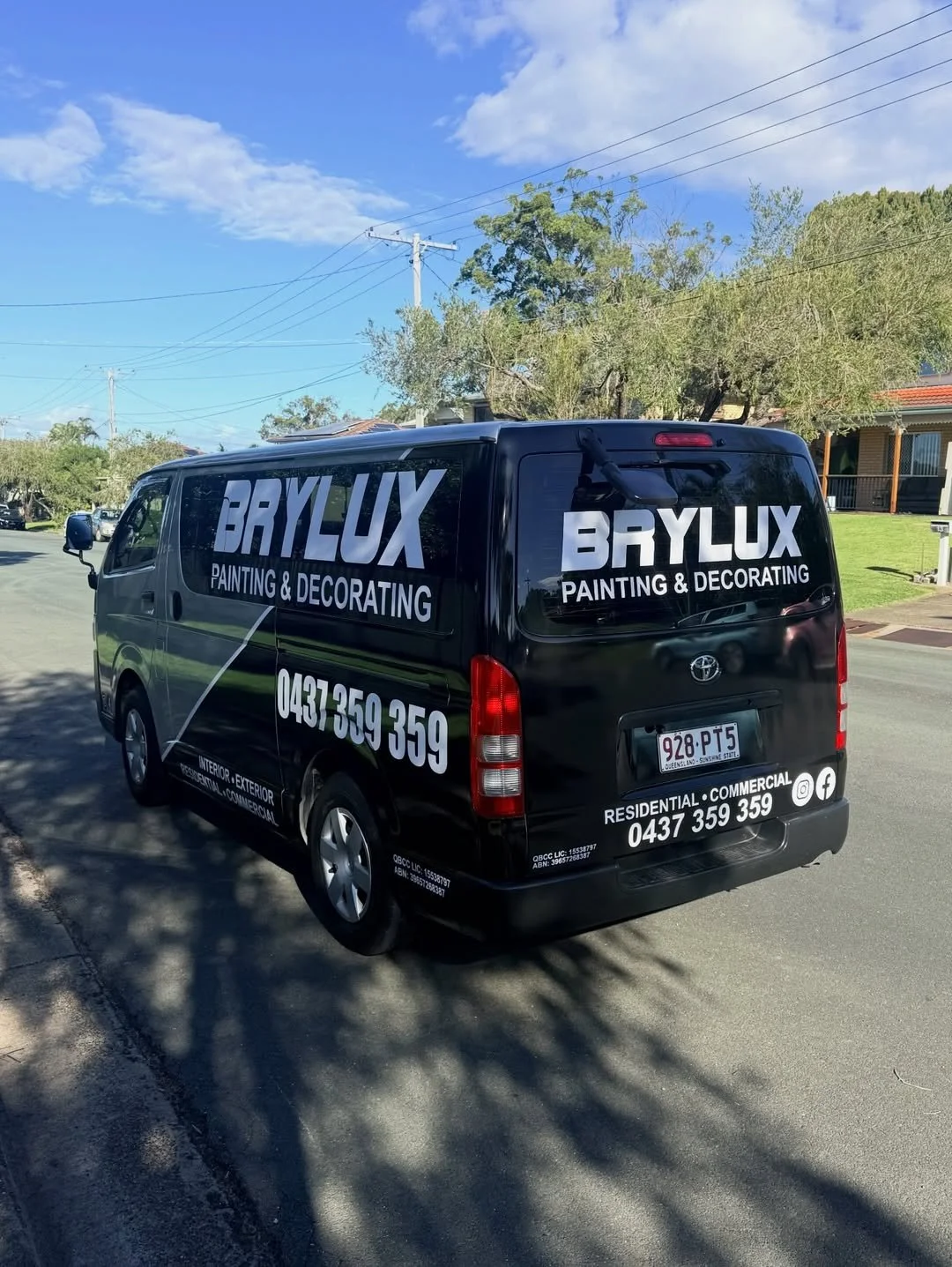 Black van with advertising for Brylux Painting and Decorating, parked on a residential street with trees and houses, under a partly cloudy sky.