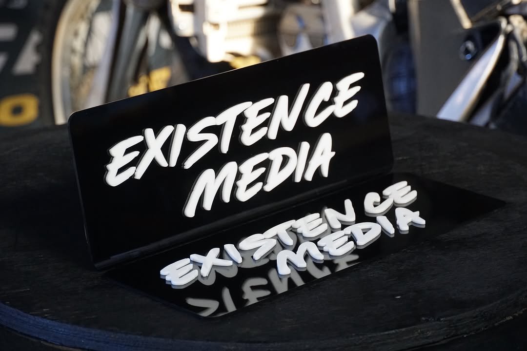 A black sign with white 3D lettering that reads 'EXISTENCE MEDIA,' placed on a black surface, with a mirrored reflection of the sign visible below.