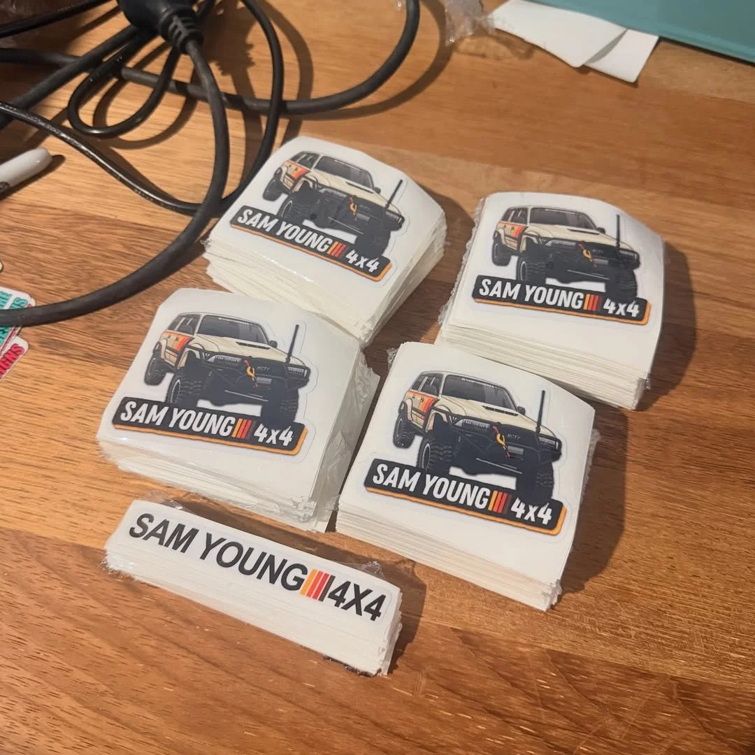 Stacks of stickers with a vehicle illustration and the text "SAM YOUNG 4X4" on a wooden surface, with some electrical cords nearby.