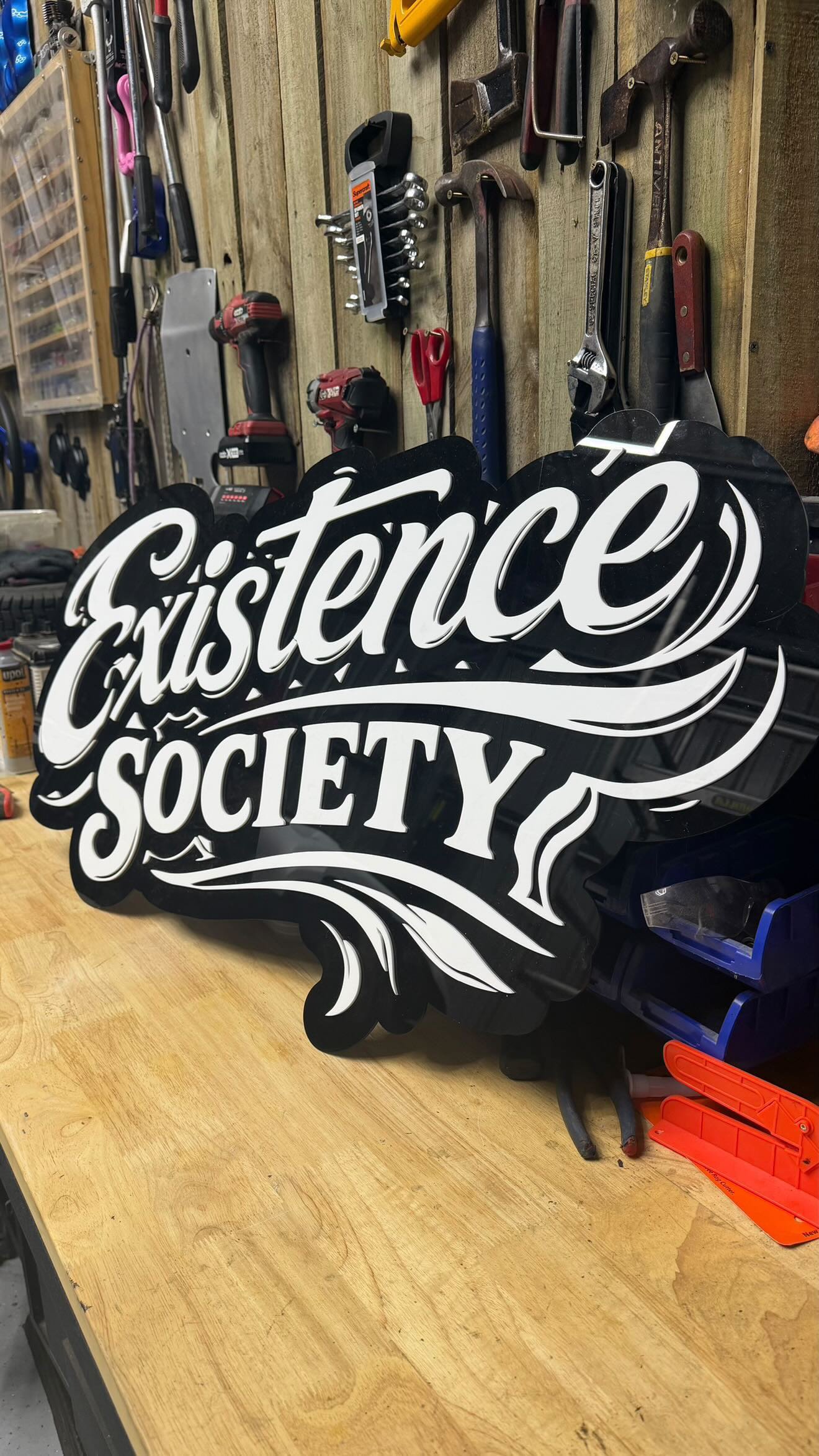 A black and white sign that reads 'Sistence Society' leaning against a workbench in a woodworking or hardware workshop. The background shows various tools hung on a wooden wall, including clamps, screwdrivers, wrenches, and a cordless drill.