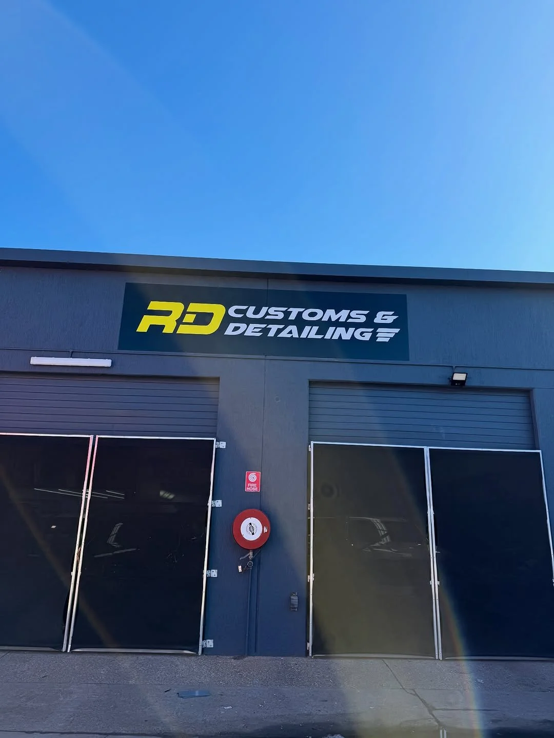 Building with a sign that reads R&D Customs & Detailing, with two garage doors and a fire hose reel on the wall