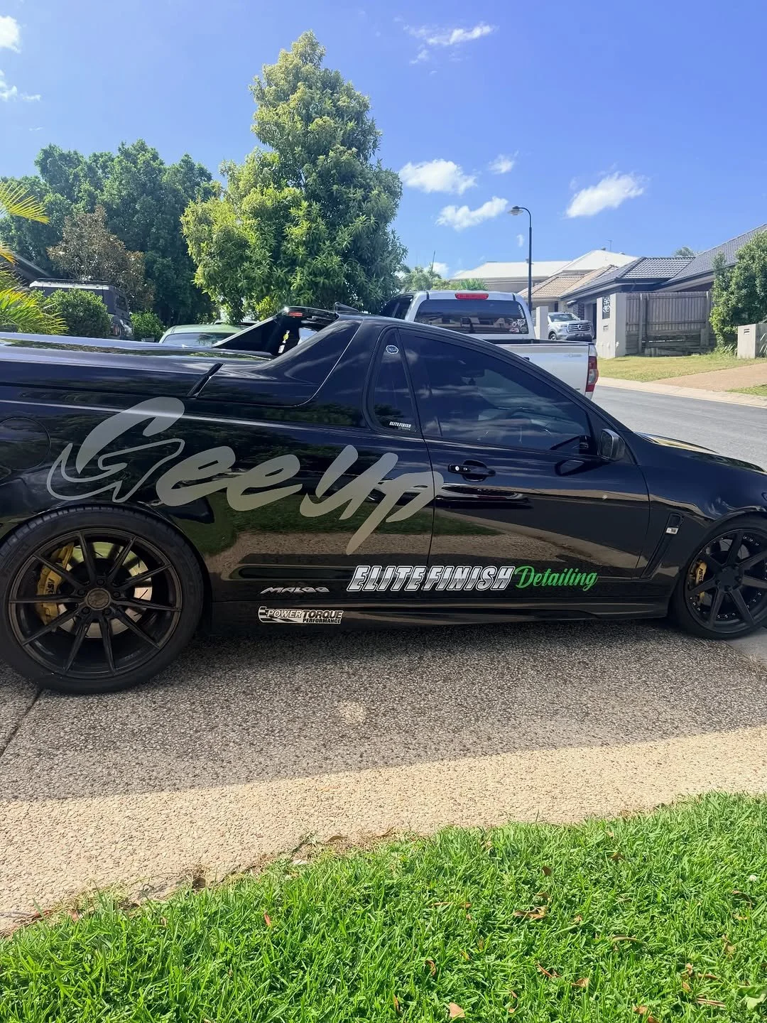 A black sports car with decals and custom wheels parked on a residential street under a clear blue sky.