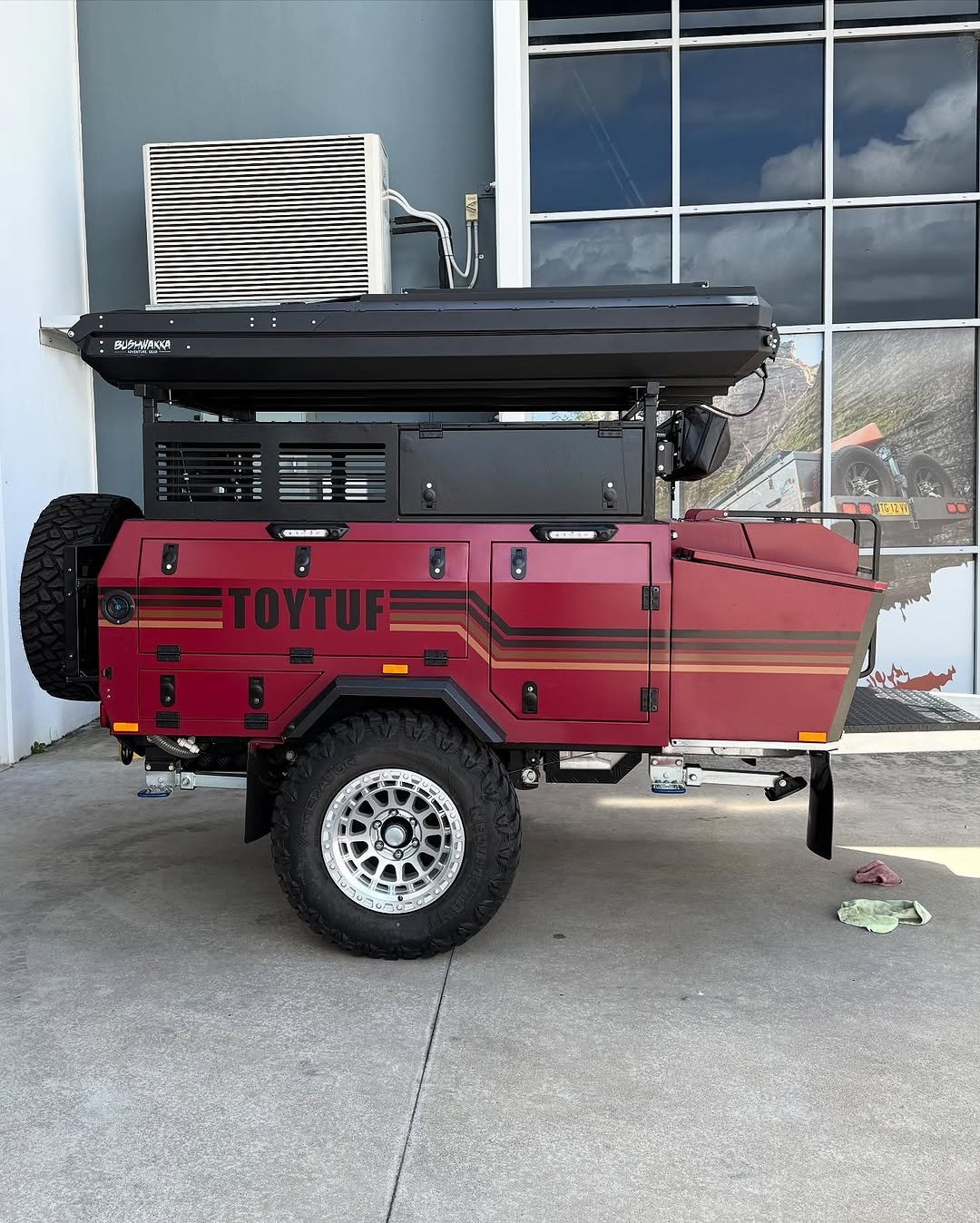 A red off-road camping trailer with large black tires, labeled "TOYTUFF", parked on a concrete surface outside a building with large glass windows, and an air conditioning unit mounted on the wall.