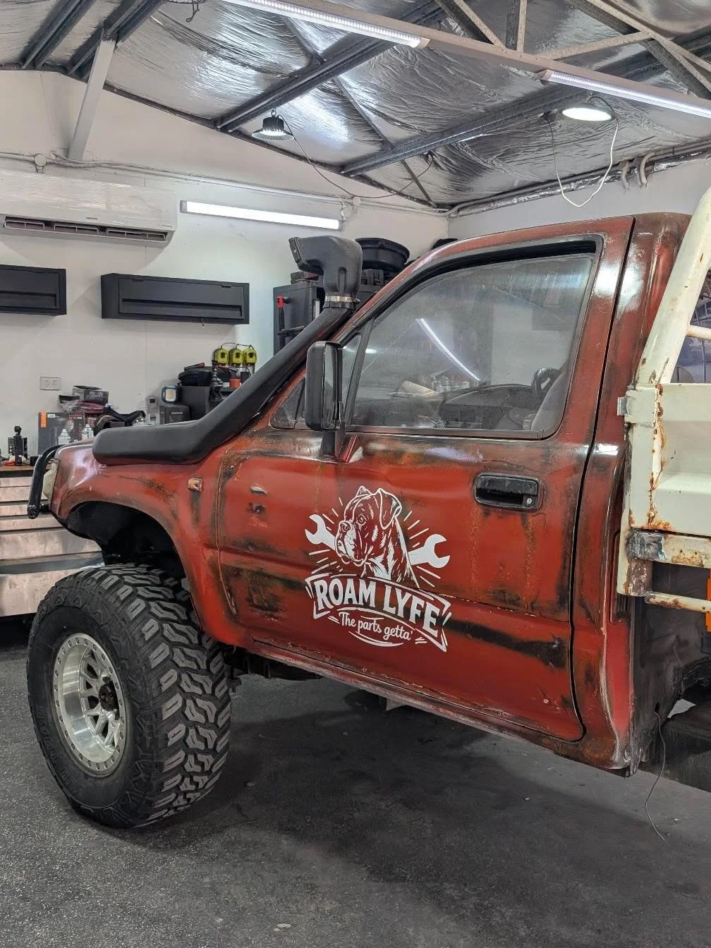 A rusty red pickup truck with a white graphic of a dog and tools, and the text 'ROAM LYFE' and 'The parts getta', inside a garage or workshop.