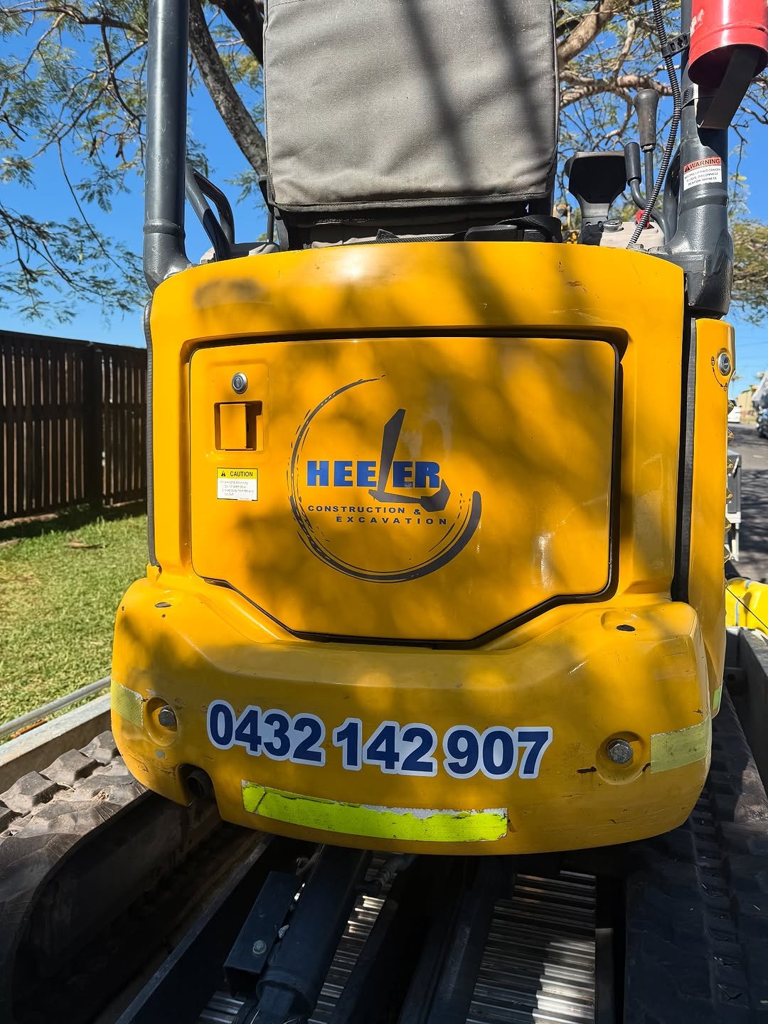 Close-up of a yellow construction and excavation vehicle with a company logo, phone number, and reflector on the back, outdoors with trees and a wooden fence.