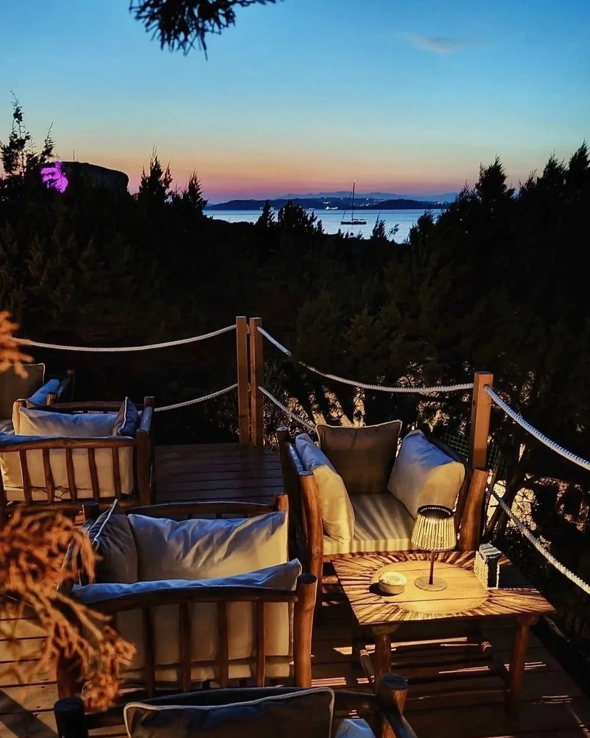 La terrazza del Puntamar a Baja Sardinia con divani di legno e cuscini, vista sul mare al tramonto, con barca a vela in mare e un paesaggio serale con cielo colorato.