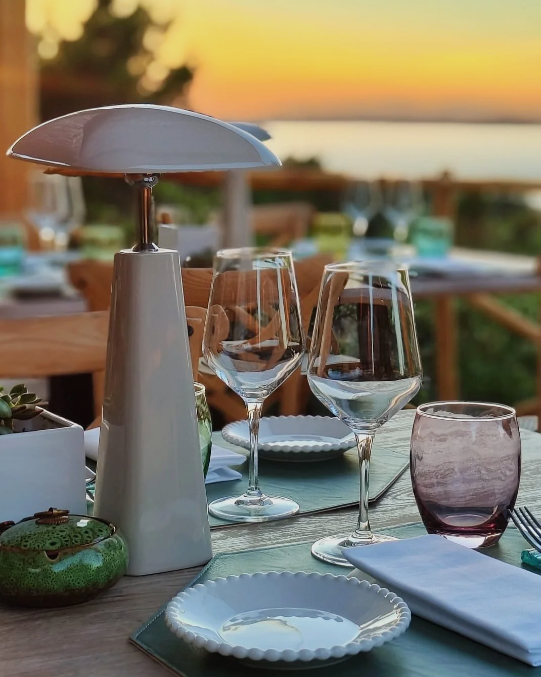 Tavola apparecchiata sulla terrazza del Puntamar a Baja Sardinia con bicchieri di vino, piatti di ceramica, un piattino, tovaglioli, e una lampada da tavolo, con un tramonto sul mare sullo sfondo.