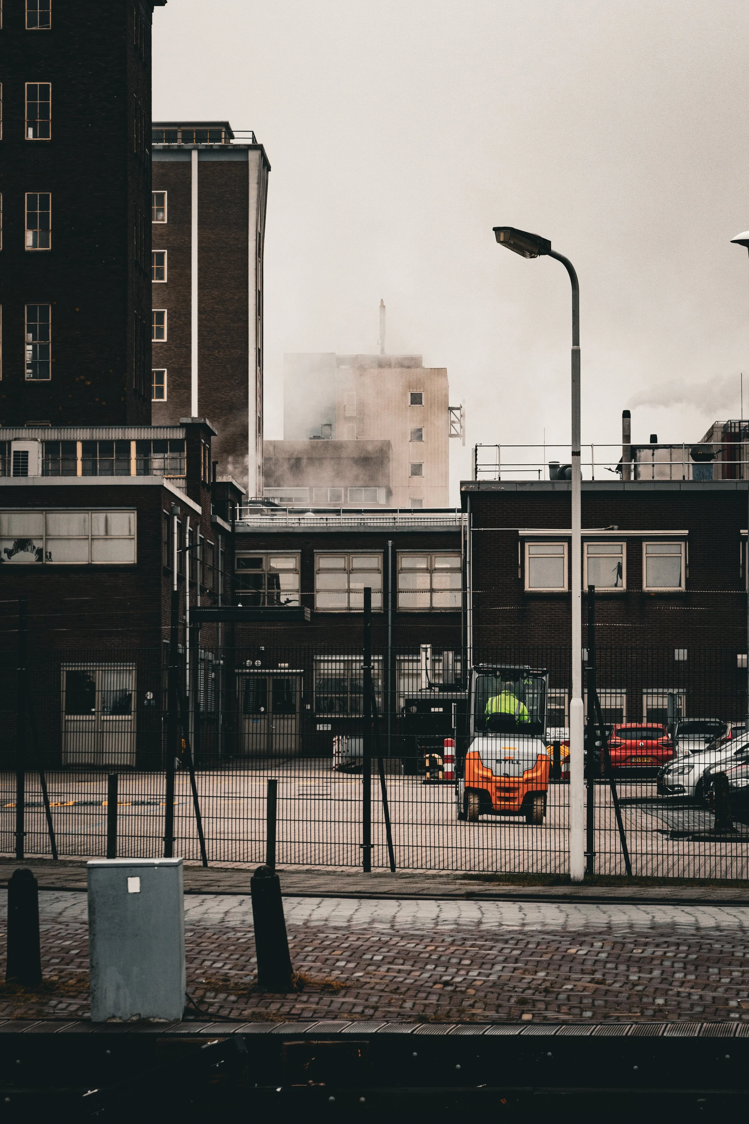 Stadskantoor of bedrijfsgebouw met rechtopstaande ramen, omgeven door een hek, met een parkeerterrein en een vrachtwagen en enkele auto's, en een gebouw dat uit de mist opkomt op de achtergrond.