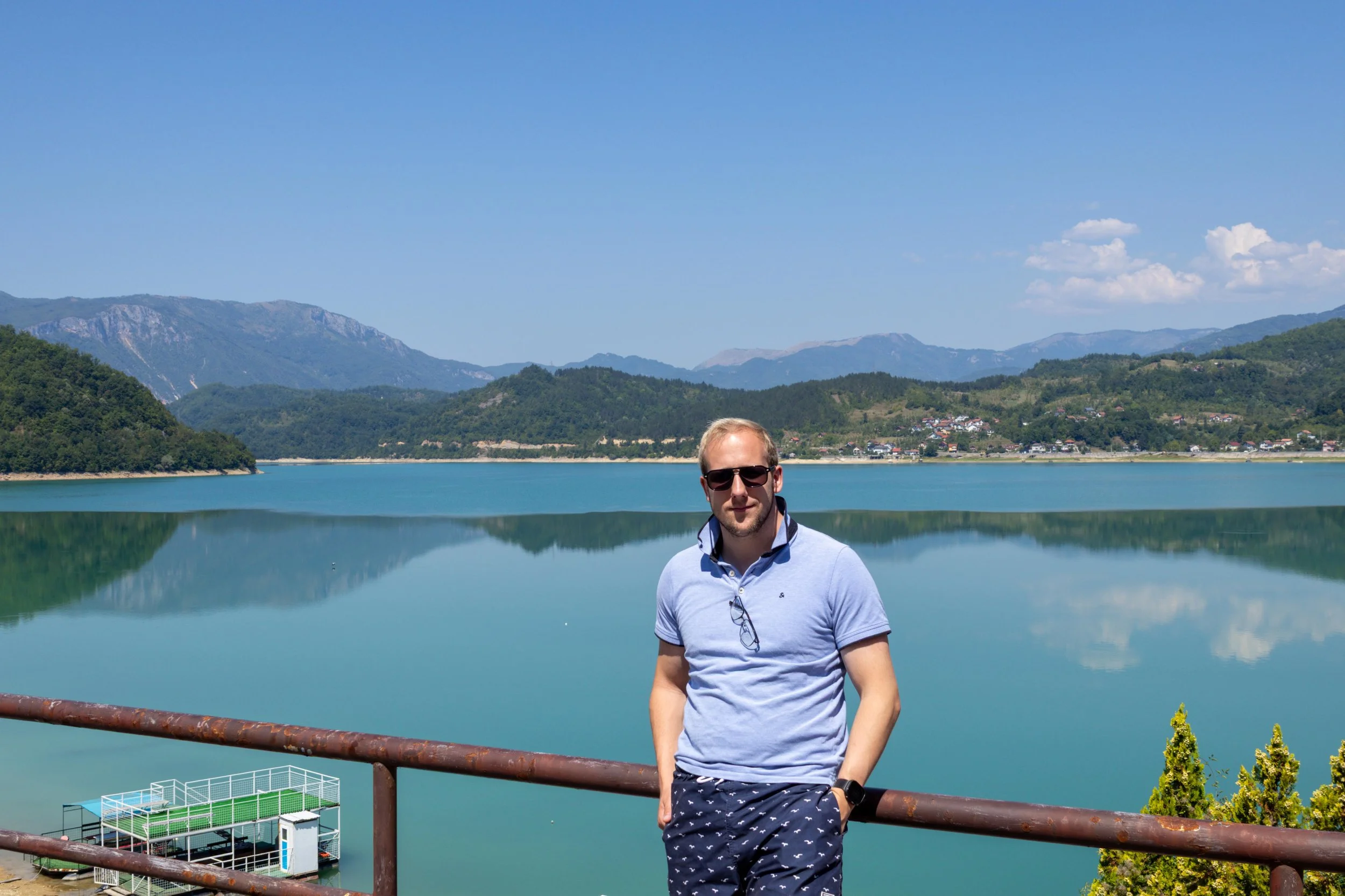 Man met zonnebril en kraagje, staand voor een meer met bergen op de achtergrond, heldere blauwe lucht en enkele wolken.