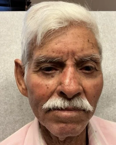 Close-up of an elderly man with white hair, bushy white eyebrows, a mustache, and a pink shirt, sitting in front of a neutral background.