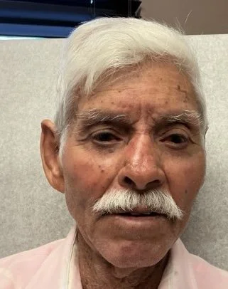 Close-up of an elderly man with white hair and a mustache, wearing a light pink shirt.