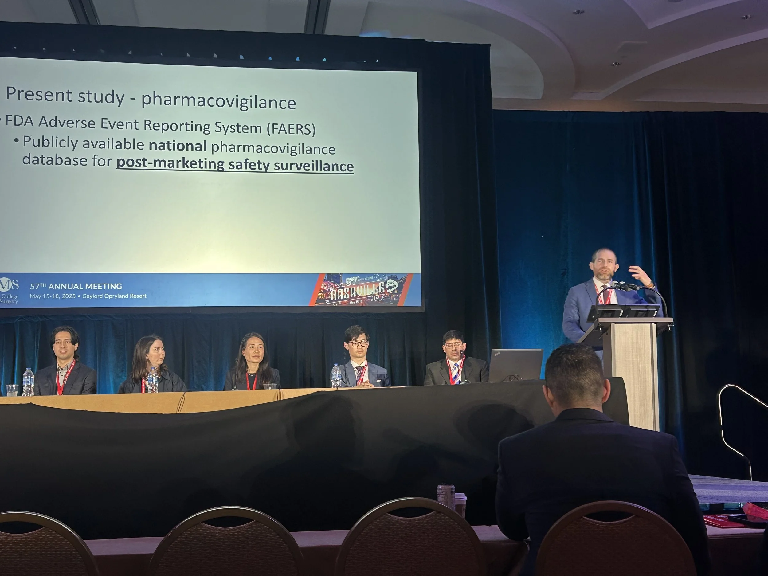 A conference room with a large presentation screen displaying a slide about pharmacovigilance and the FDA Adverse Event Reporting System, with a panel of five people sitting at a table and one man standing at a podium.