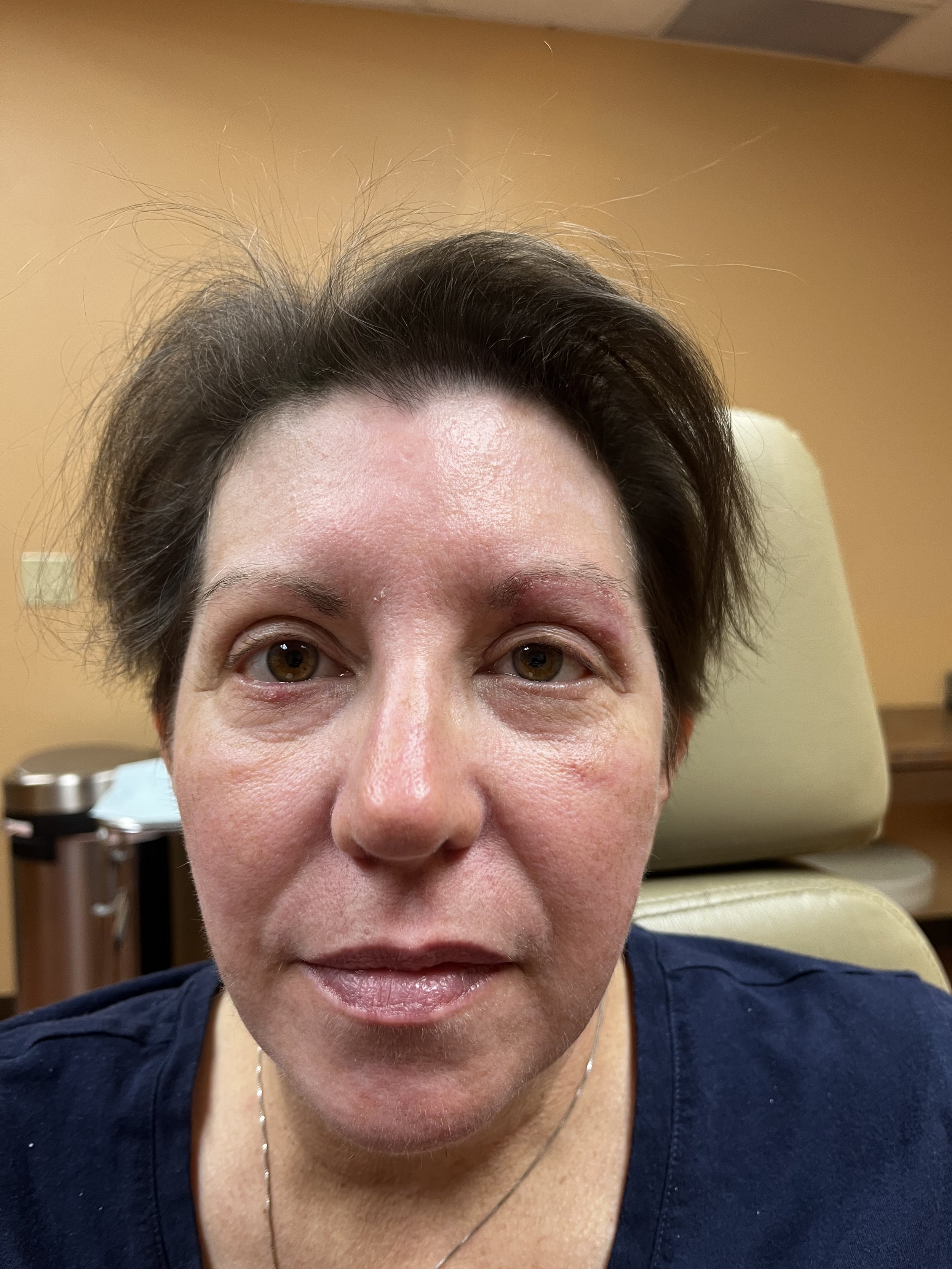 A close-up of a woman with short dark hair, brown eyes, and light skin. She is wearing a navy shirt and a necklace. She appears to be sitting in a medical or examination room with beige walls and medical equipment in the background.