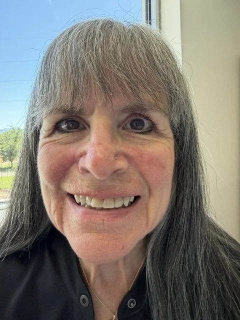 Close-up of a smiling woman with long gray hair, blue eyes, and a nose piercing, standing indoors near a window with a view of trees and blue sky.