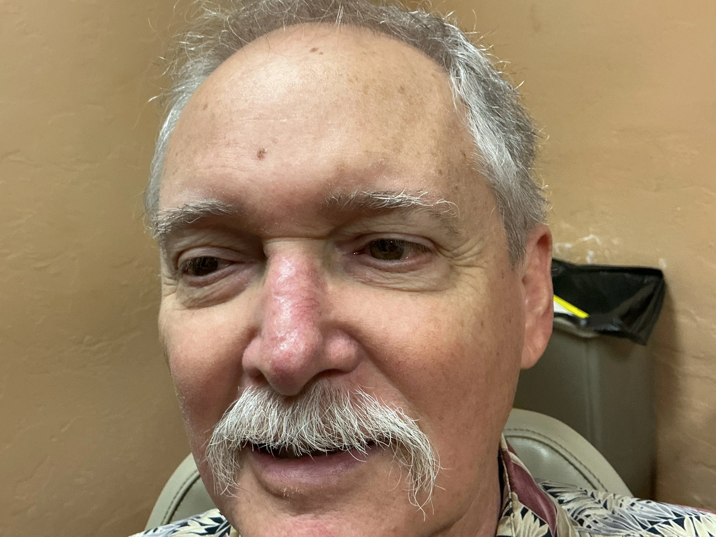 Close-up of an elderly man with gray hair and a beard, smiling slightly, with a beige wall in the background.