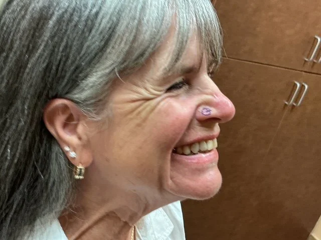 Side profile of a smiling older woman with gray hair, earrings, and a noseband.