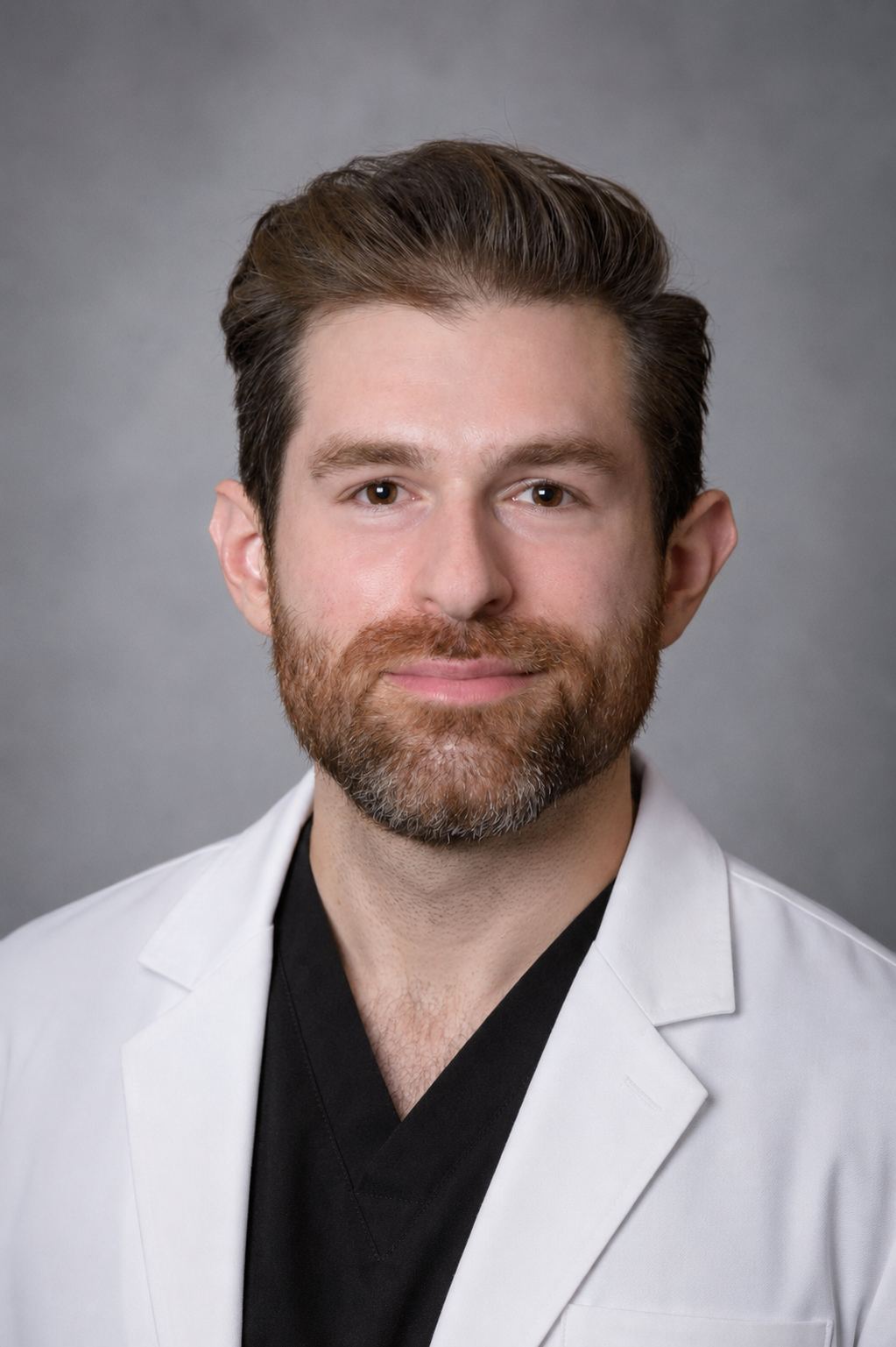 Headshot of a man with brown hair, beard, and brown eyes, wearing a white coat over black clothing, against a gray background.