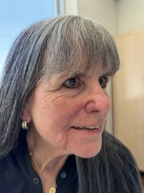Close-up of a middle-aged woman with long gray hair and bangs, wearing an earring and a necklace, smiling slightly, indoors near a window.