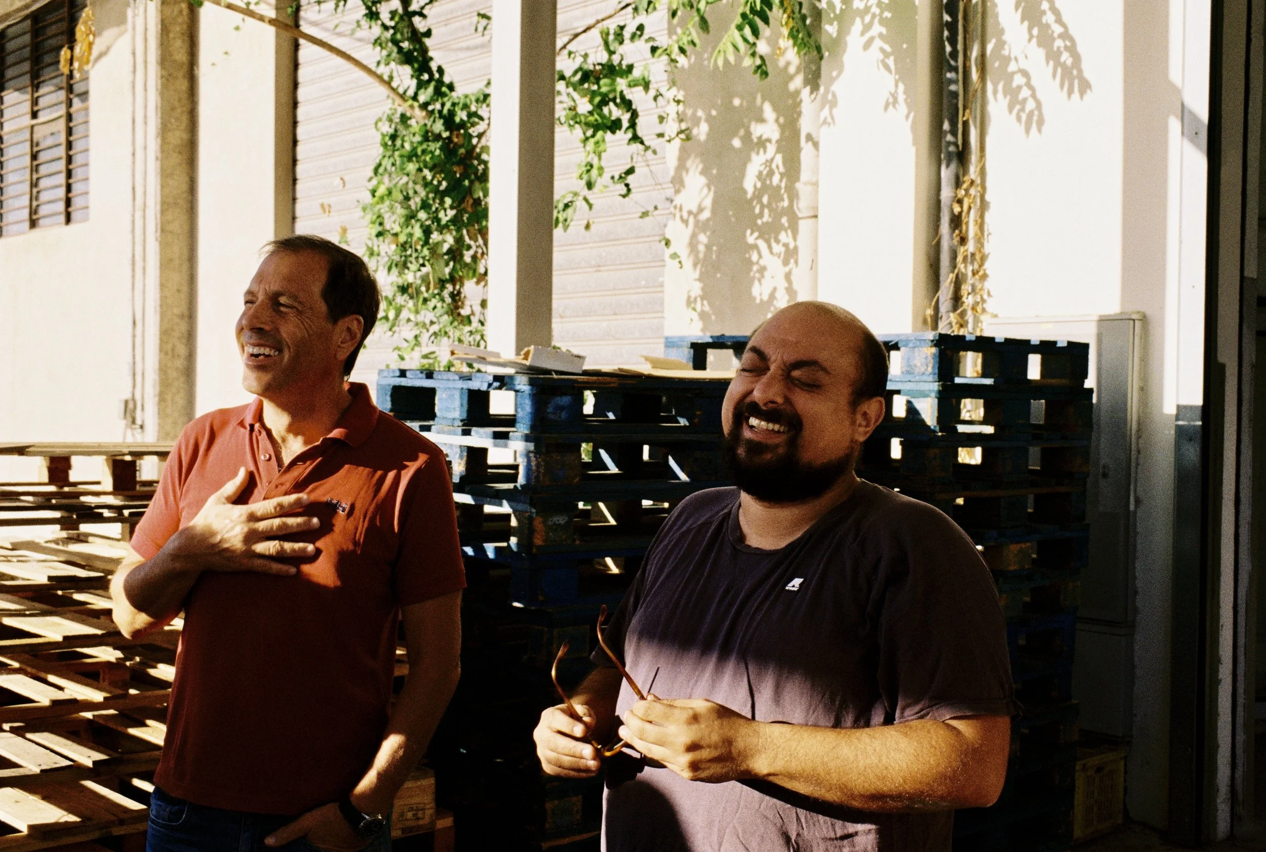 Twee mannen lachen en praten terwijl ze buiten staan, omgeven door pallets en een muur met planten en schaduw.