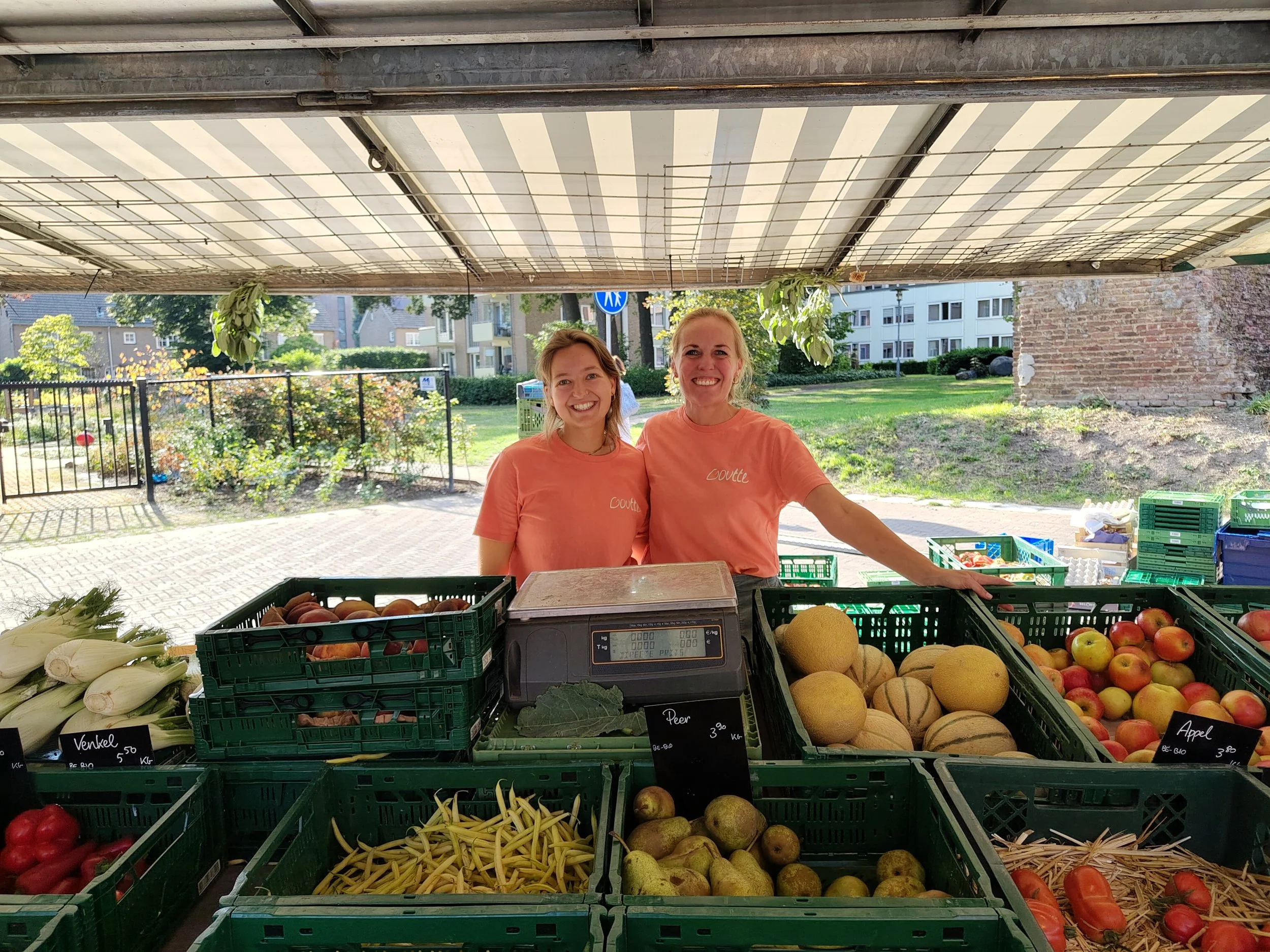 Twee vrouwen in oranje t-shirts staan achter een fruitschaal met verschillende soorten fruit op een markt, onder een overkapping.