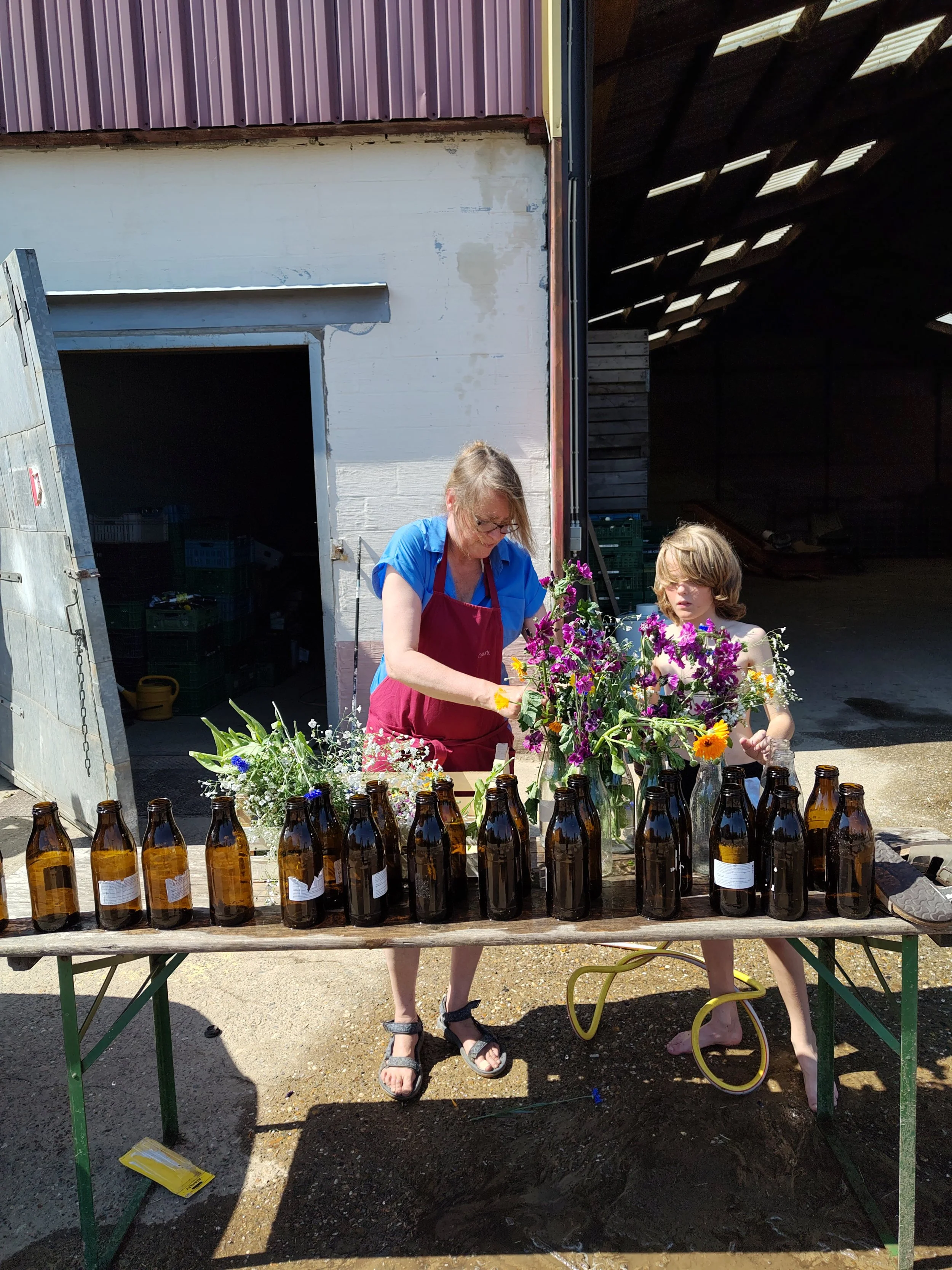 Een vrouw en een jongen staan naast een tafel met lege bierflessen en bloemen, buiten bij een stal of schuur.