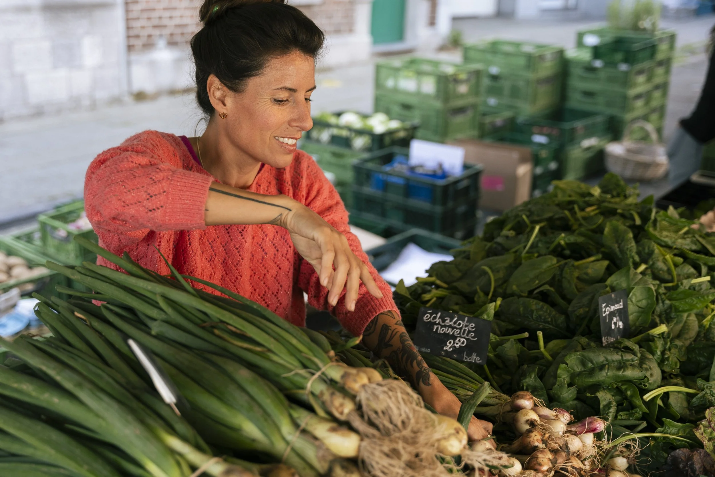 Vrouw in een markt met groene groenten en knoflook, lachend en naar beneden kijkend.
