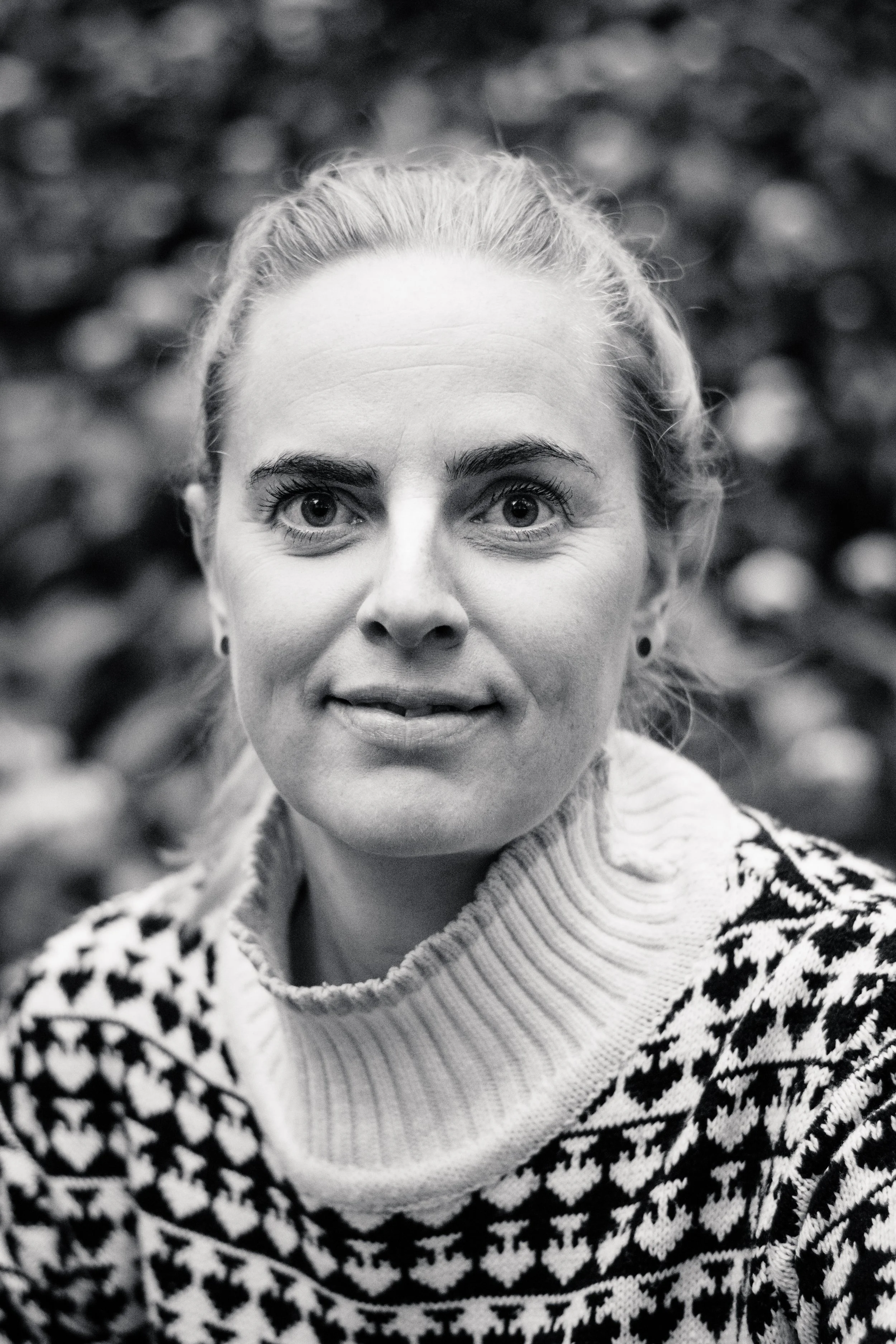 Sort-hvid portrætfotografi af en kvinde med blondt hår, iført strikket sweater med sort og hvidt mønster, foran en uklar baggrund.