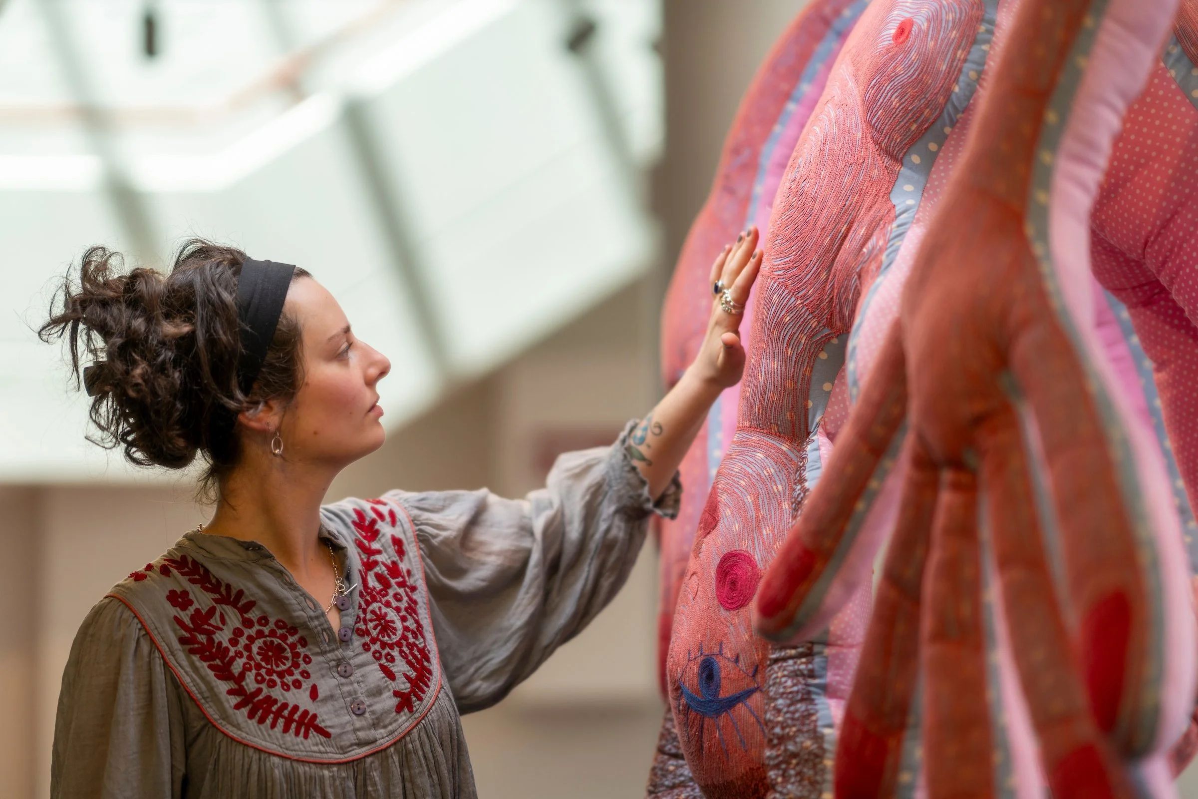 A woman with curly hair, wearing a headband and a beige shirt with red embroidery, is touching a colorful quilt hanging on a display. The quilt has various pink, red, and blue patterns and textures.