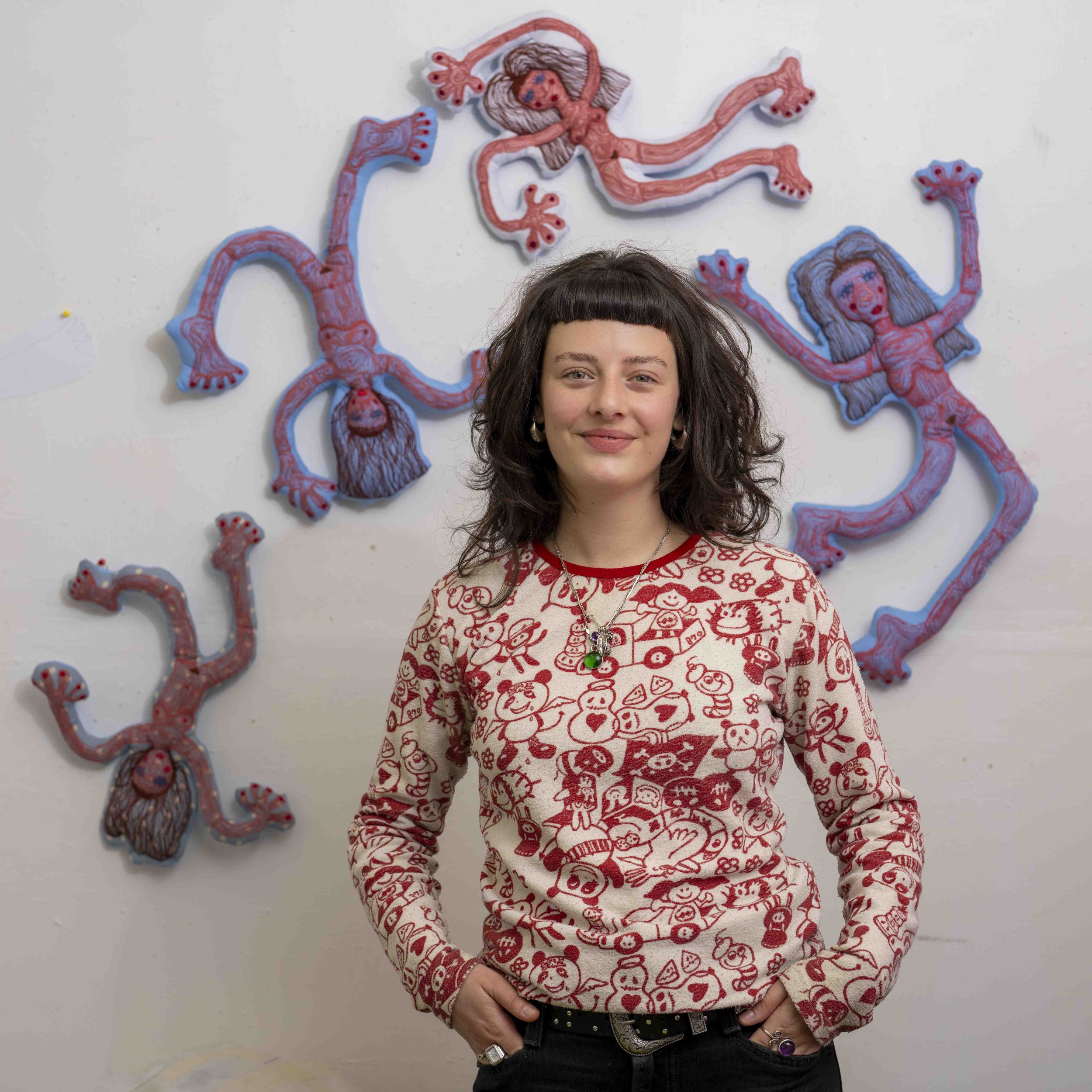 Person wearing a red sweatshirt with various cartoon faces and icons, standing in front of paper cutouts of human digestive organs on a white wall.