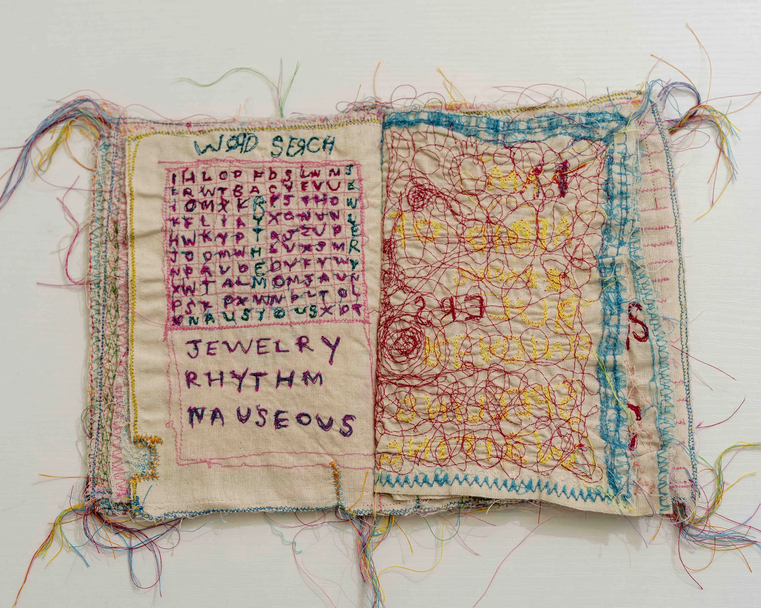 A fabric sample with various colorful embroidery stitches and wordings including 'WAD SECH,' 'JEWELRY,' and other embroidered letters, with loose threads around the edges.