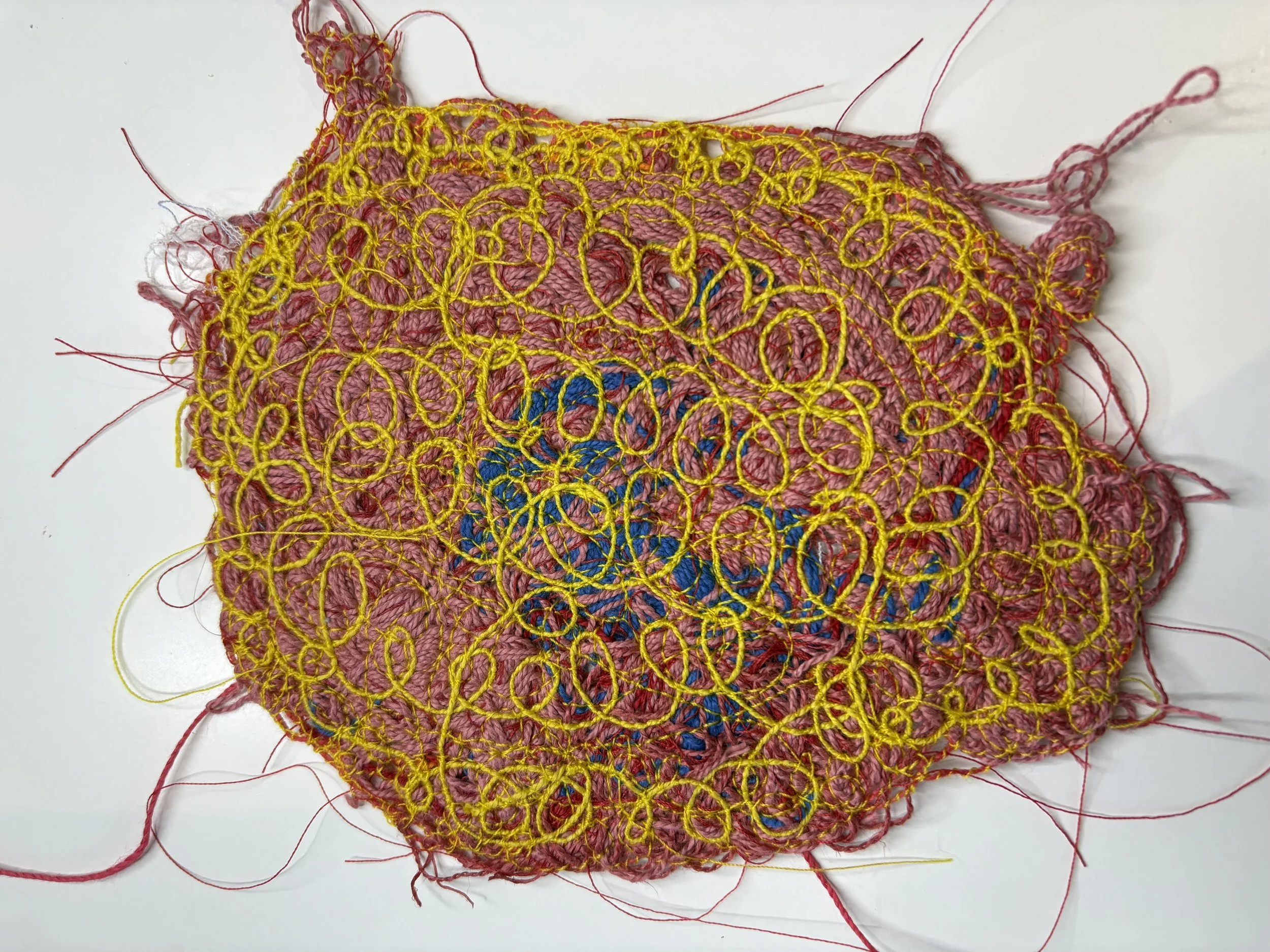 Close-up of a piece of pink woven fabric with yellow embroidery on top, featuring loops and circles. Red and pink threads extend from the fabric's edges, and there are some blue threads visible beneath the embroidery.