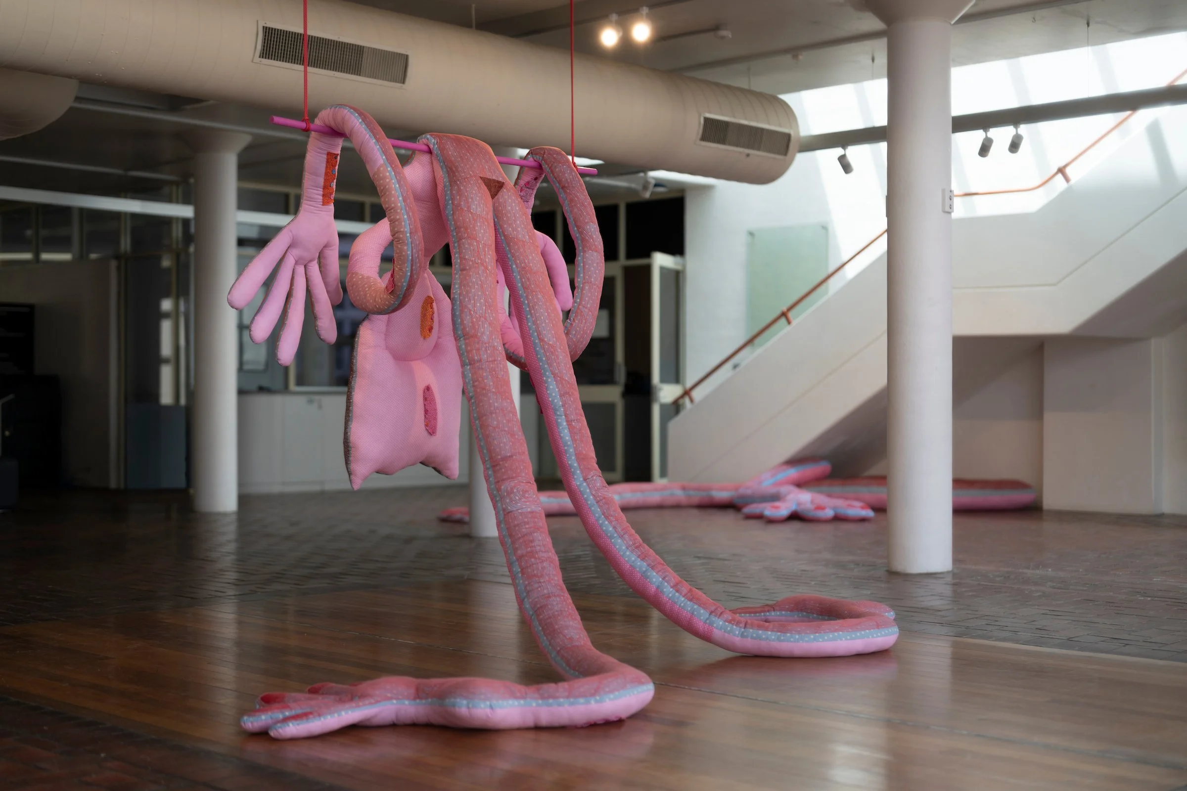 Pink plush snake sculpture with different body parts hanging from a pink pipe in a modern indoor space.