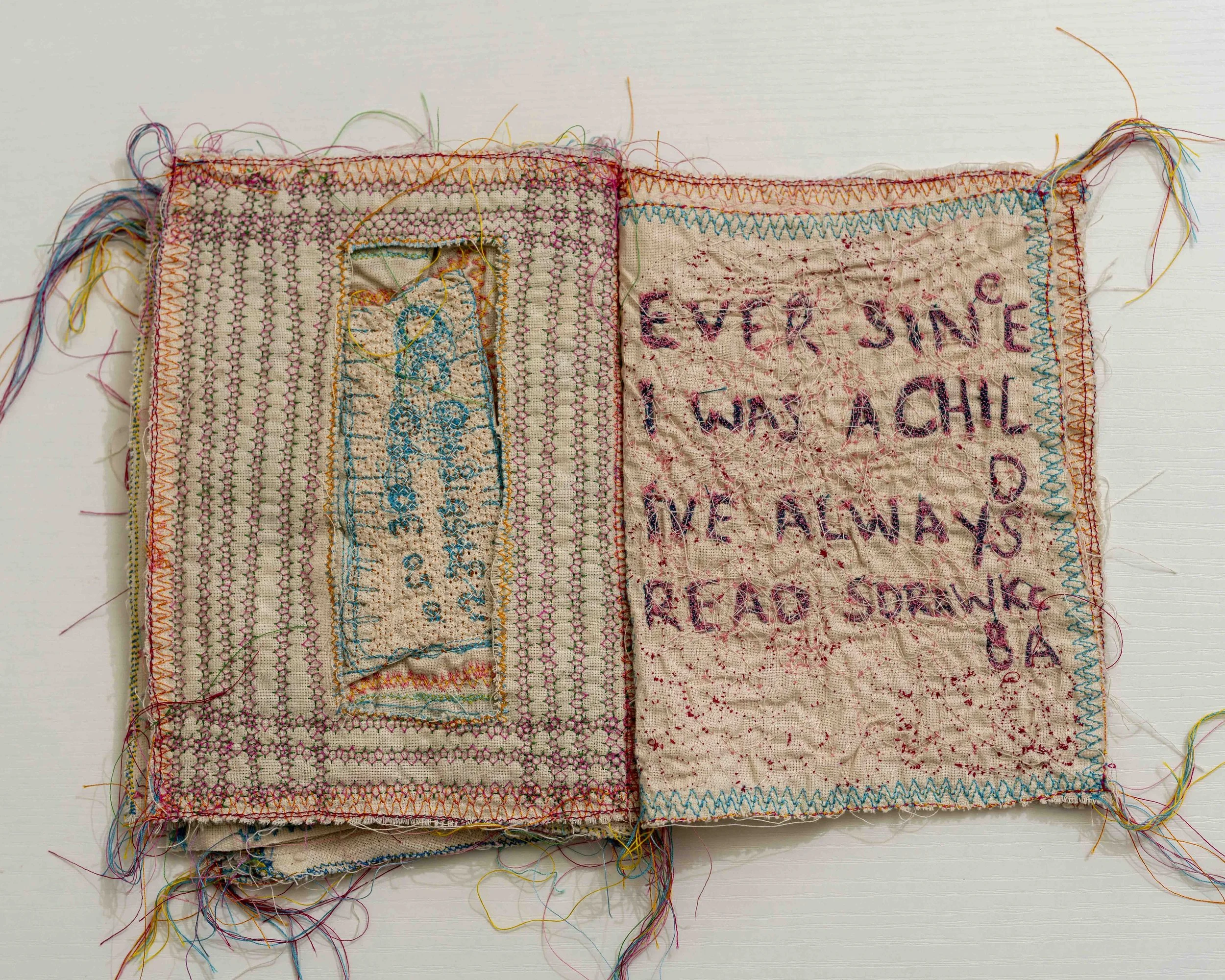 A piece of fabric with visible multicolored embroidery and loose threads, featuring stitched text and patterns.