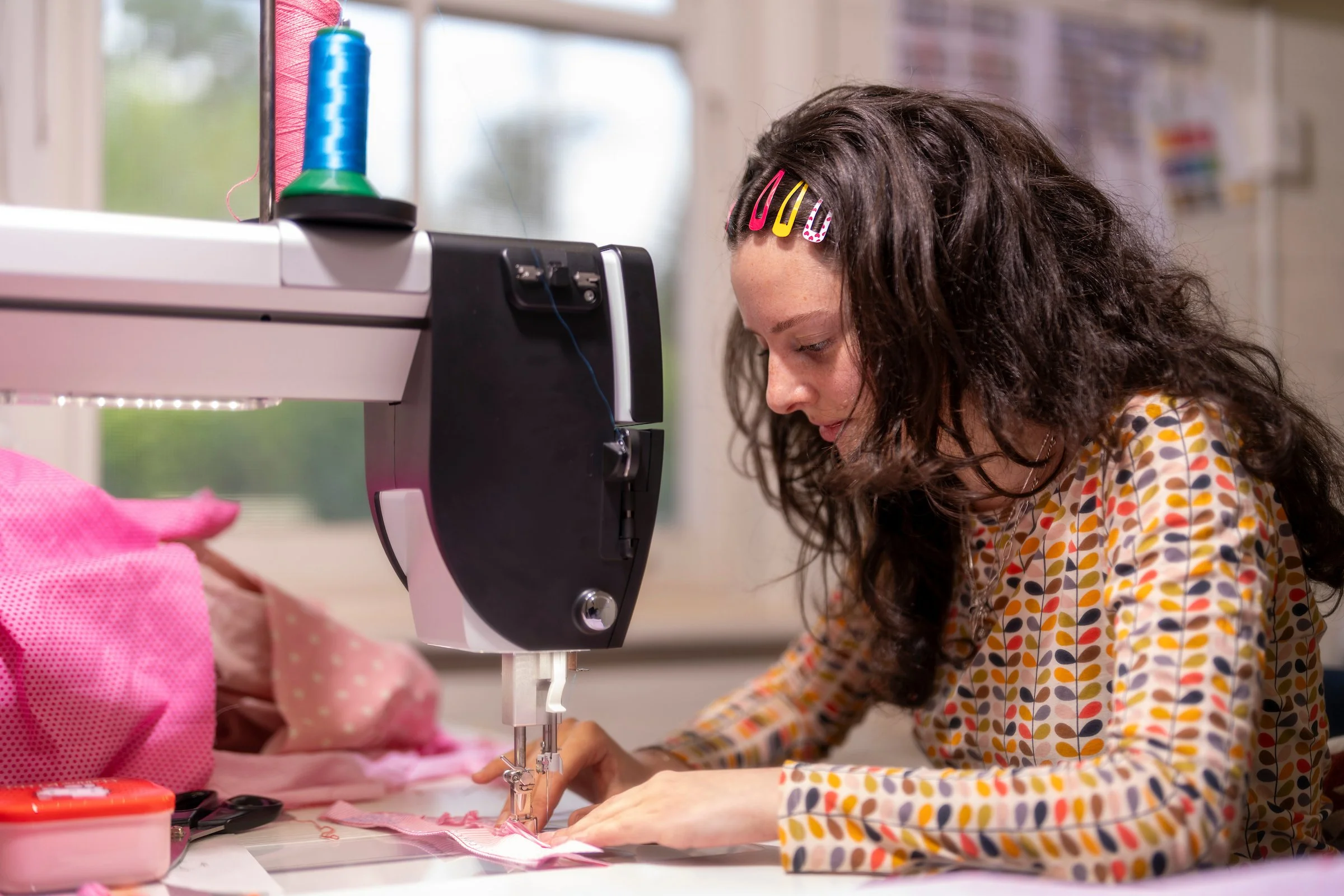 Young girl with curly hair and colorful clips in her hair, wearing a patterned dress, sewing fabric under a sewing machine in a bright room.