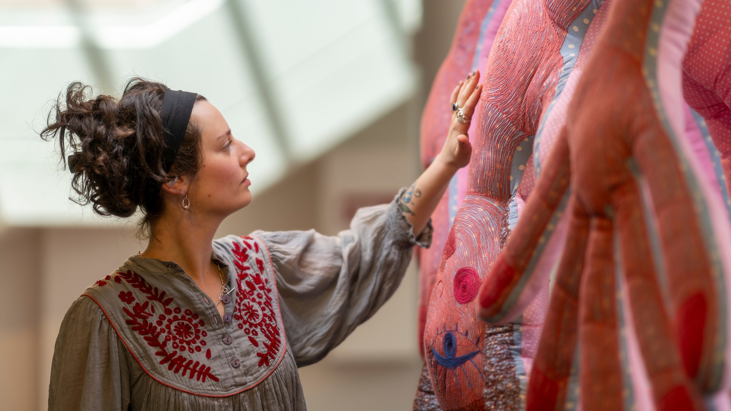 A woman with dark curly hair, wearing a black headband and a beige shirt with red embroidery, is touching a pink and orange patterned textile on a display rack.