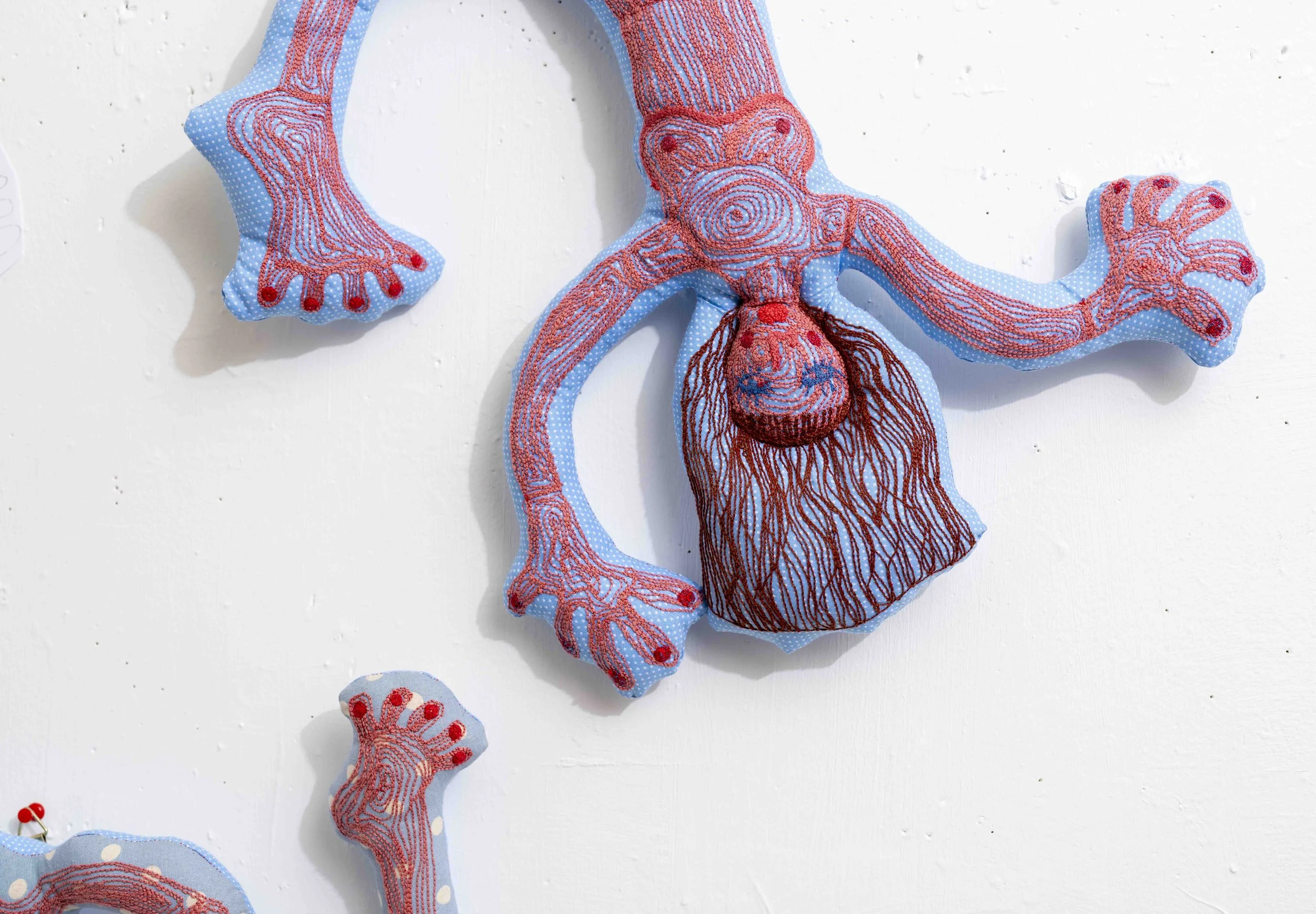 A fabric art piece resembling a stylized octopus, with an embroidered body and limbs in red and orange threads on light blue fabric with white polka dots, mounted on a white wall.