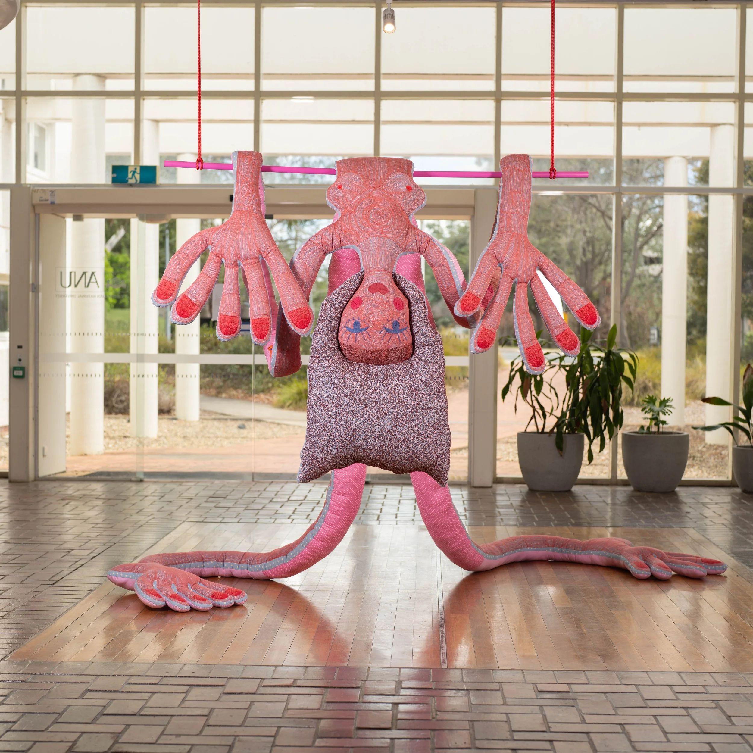 Large pink plush animal with long arms and a snake-like body, hanging from a pink bar suspended by red ropes inside a building with glass walls and plants outside.