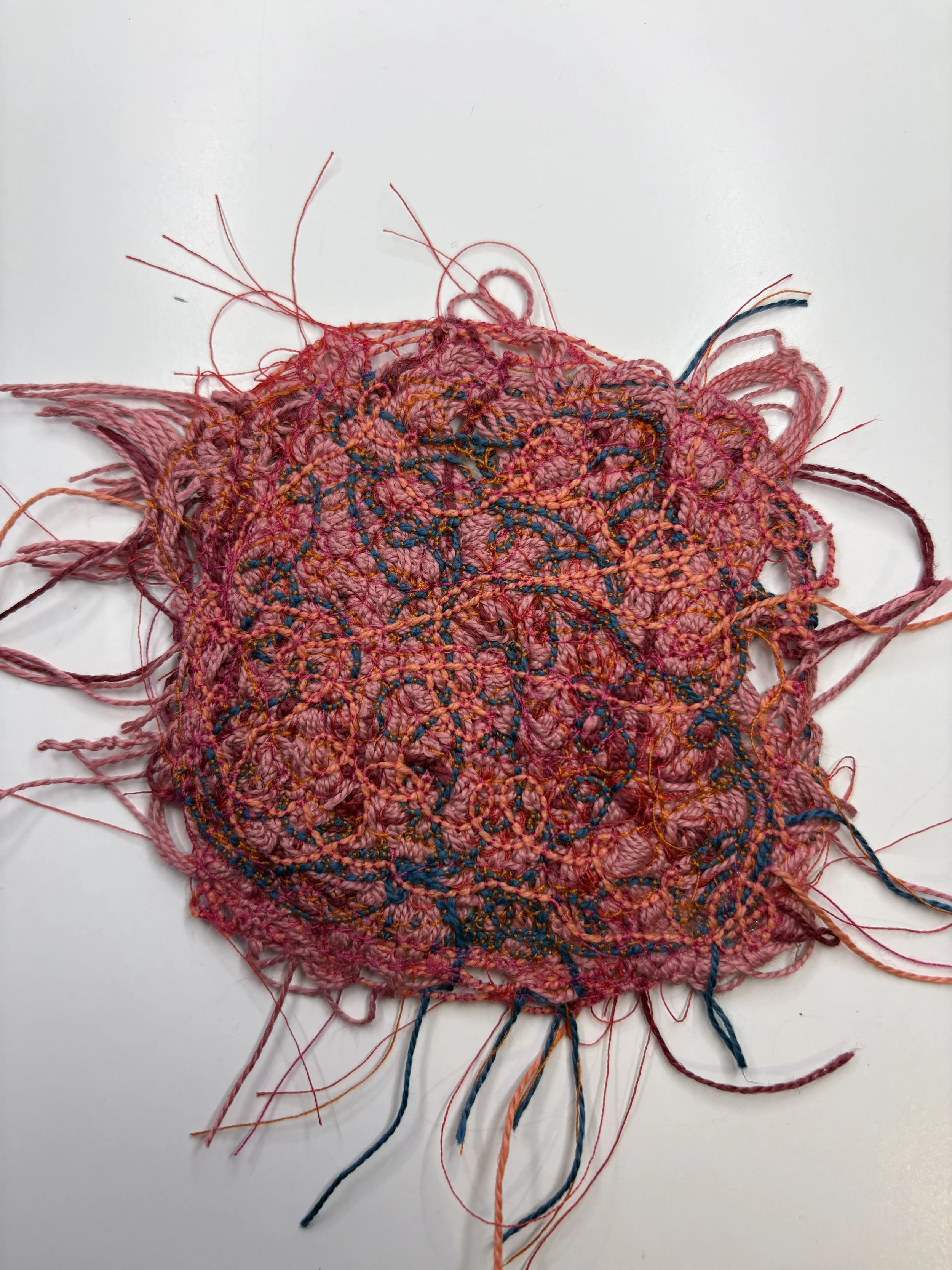 A partially finished colorful crochet piece with pink, orange, blue, and purple yarn, featuring a lacy pattern with loose ends and some strands extending outward on a white surface.