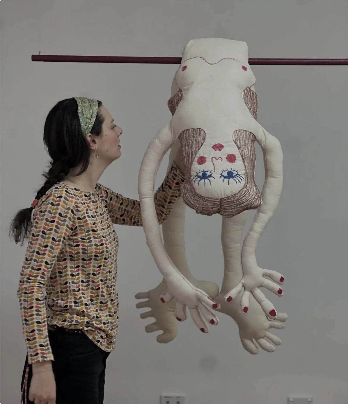 A woman with a colorful patterned shirt and a green headband is holding a large plush puppet with a human face and long limbs, hanging upside down from a pink pole.