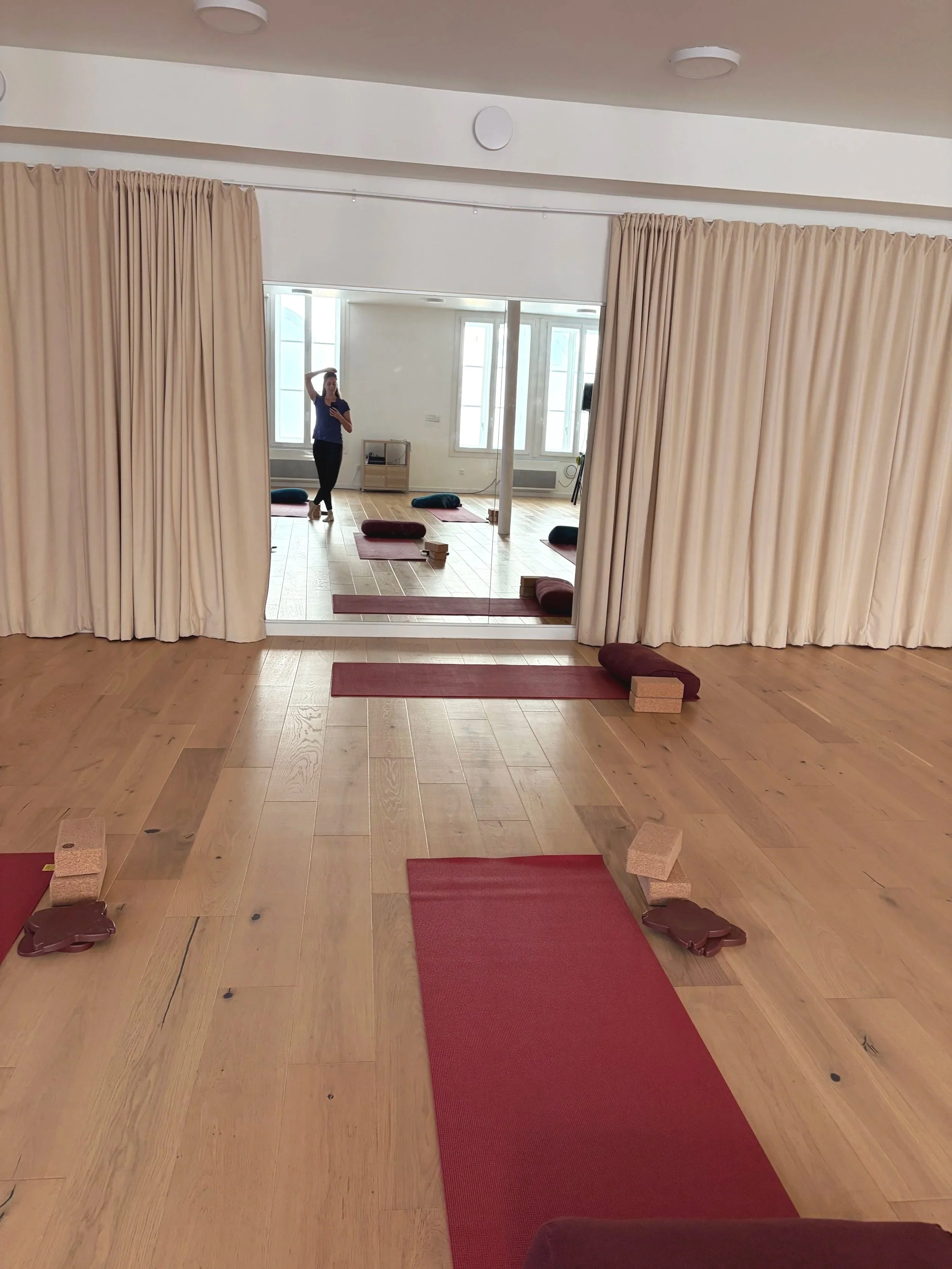 Salle de yoga avec tapis rouges et coussins sur un sol en bois clair, vue d'un miroir avec une femme prenant une photo, grande fenêtre laissant entrer beaucoup de lumière naturelle.
