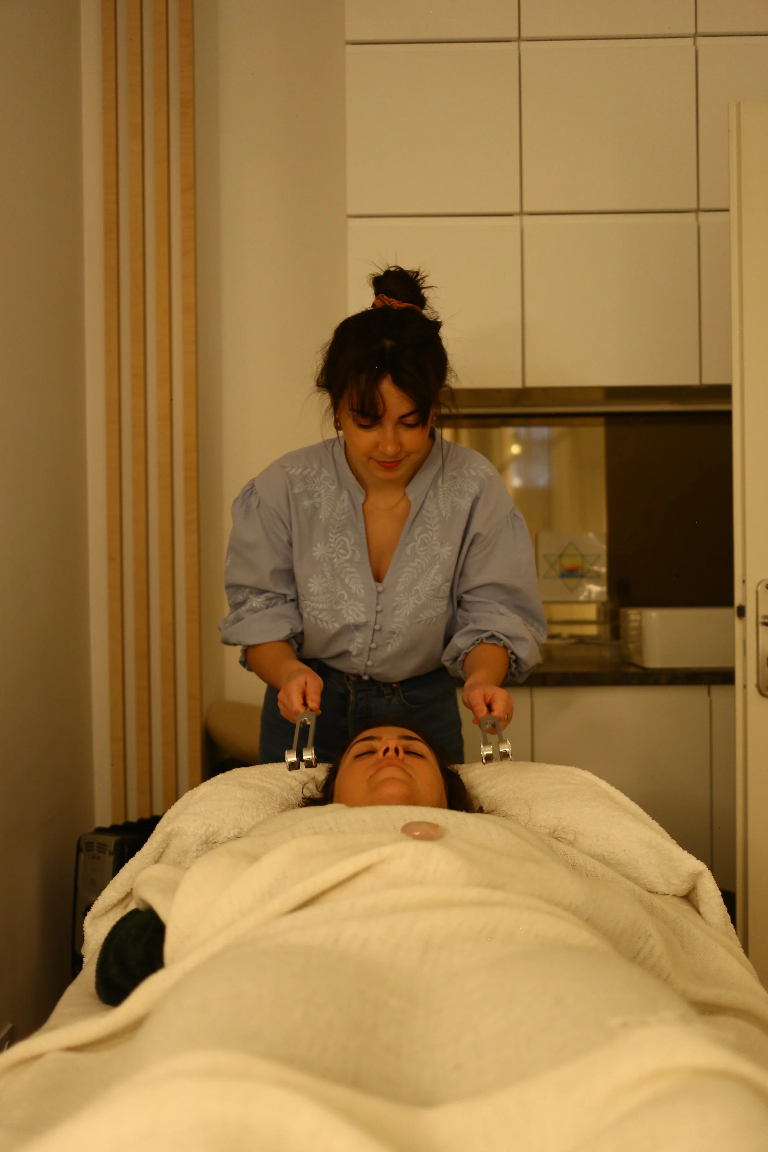 Une femme allongée sur une table d'examen médical avec un drap blanc, une autre femme effectue une procédure médicale avec des appareils métalliques. La scène se déroule dans une salle clinique ou un centre de soins.