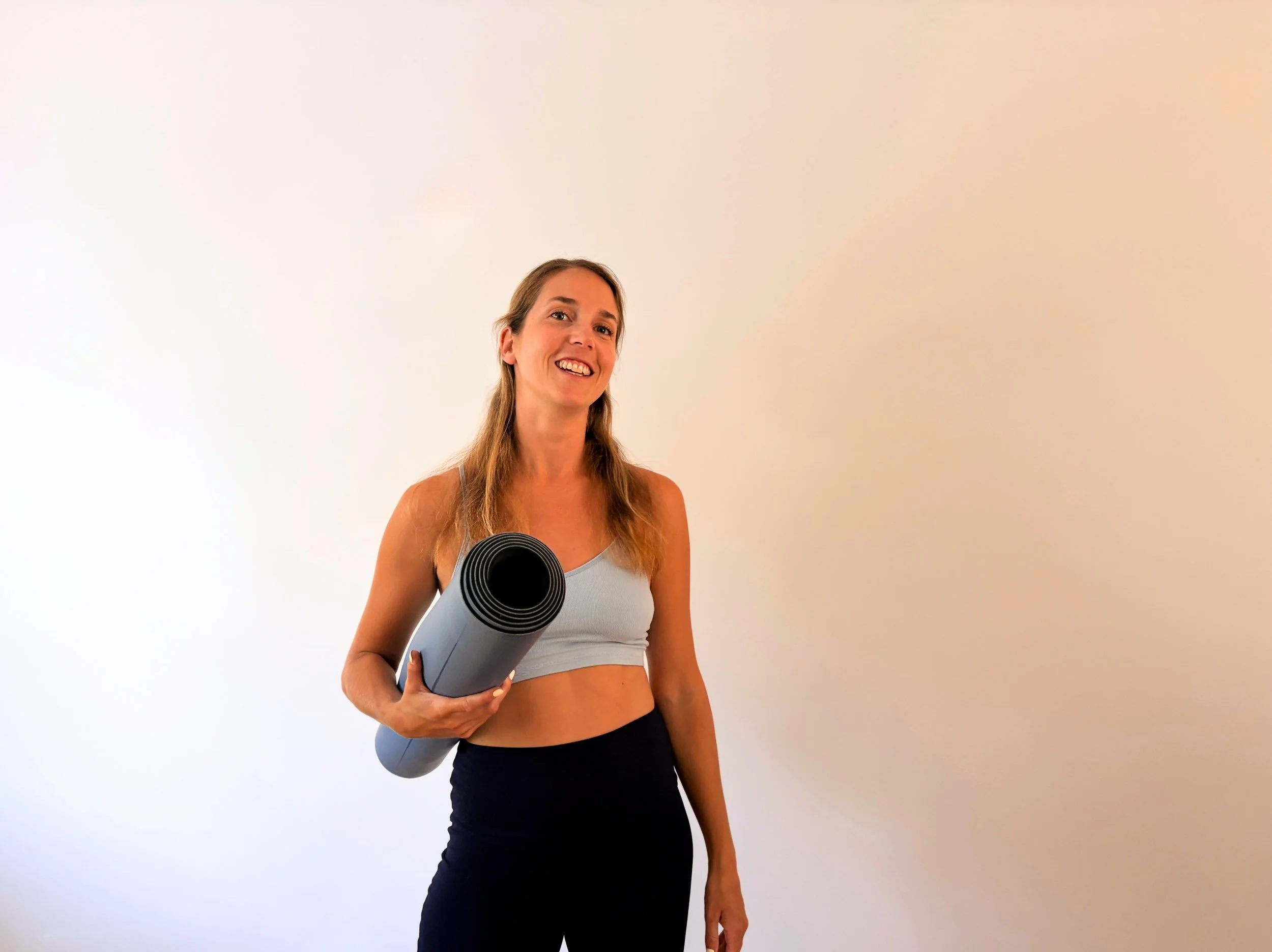 Une femme souriante portant un haut de sport gris clair et un legging noir, tenant un tapis de yoga gris enroulé sur l'épaule, debout devant un fond blanc.