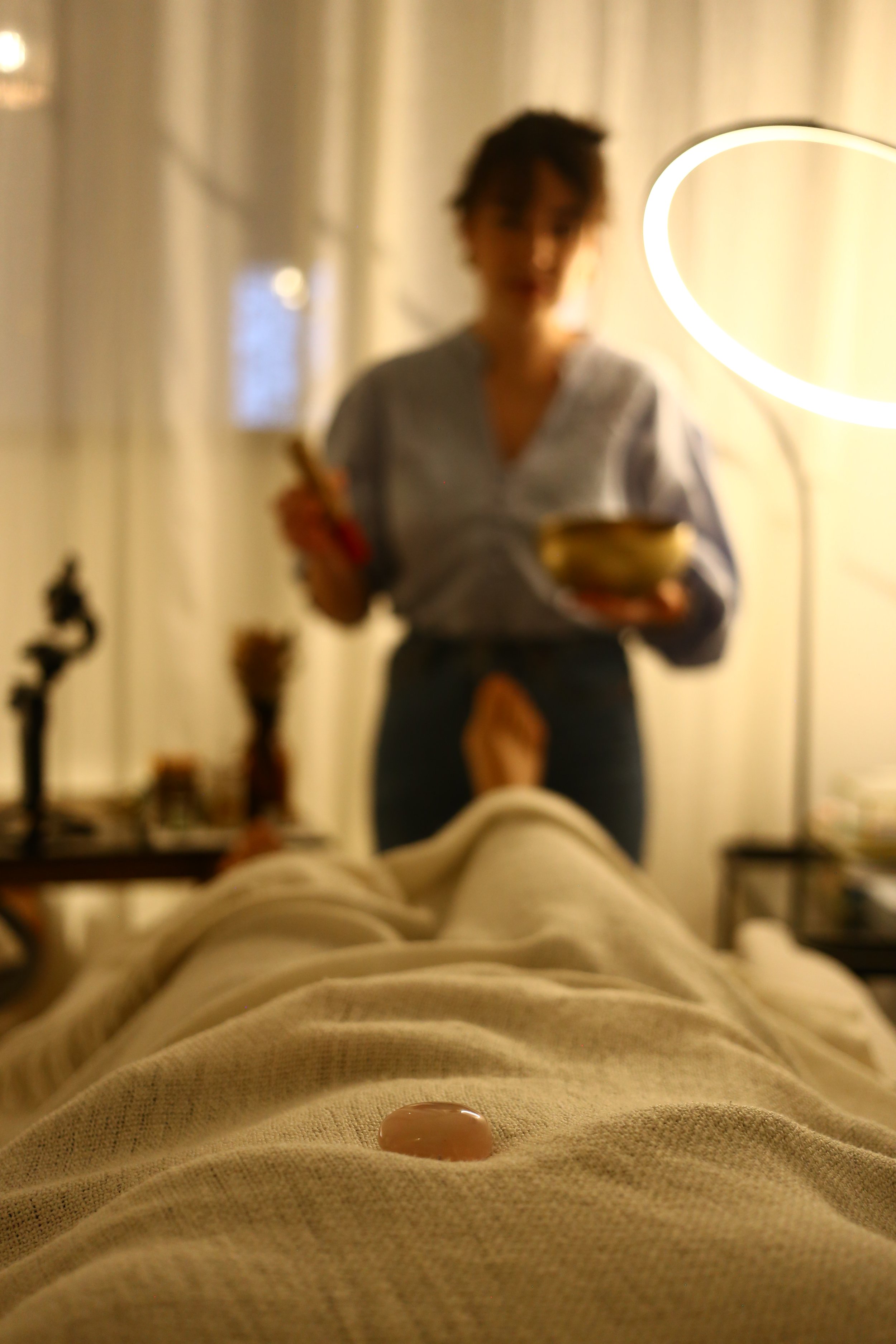 Une personne allongée sur un lit regarde vers le haut, sans vêtements, avec une substance blanche sur le ventre. En arrière-plan, une femme debout, tenant un bol et une cuillère, dans une pièce chaleureuse.
