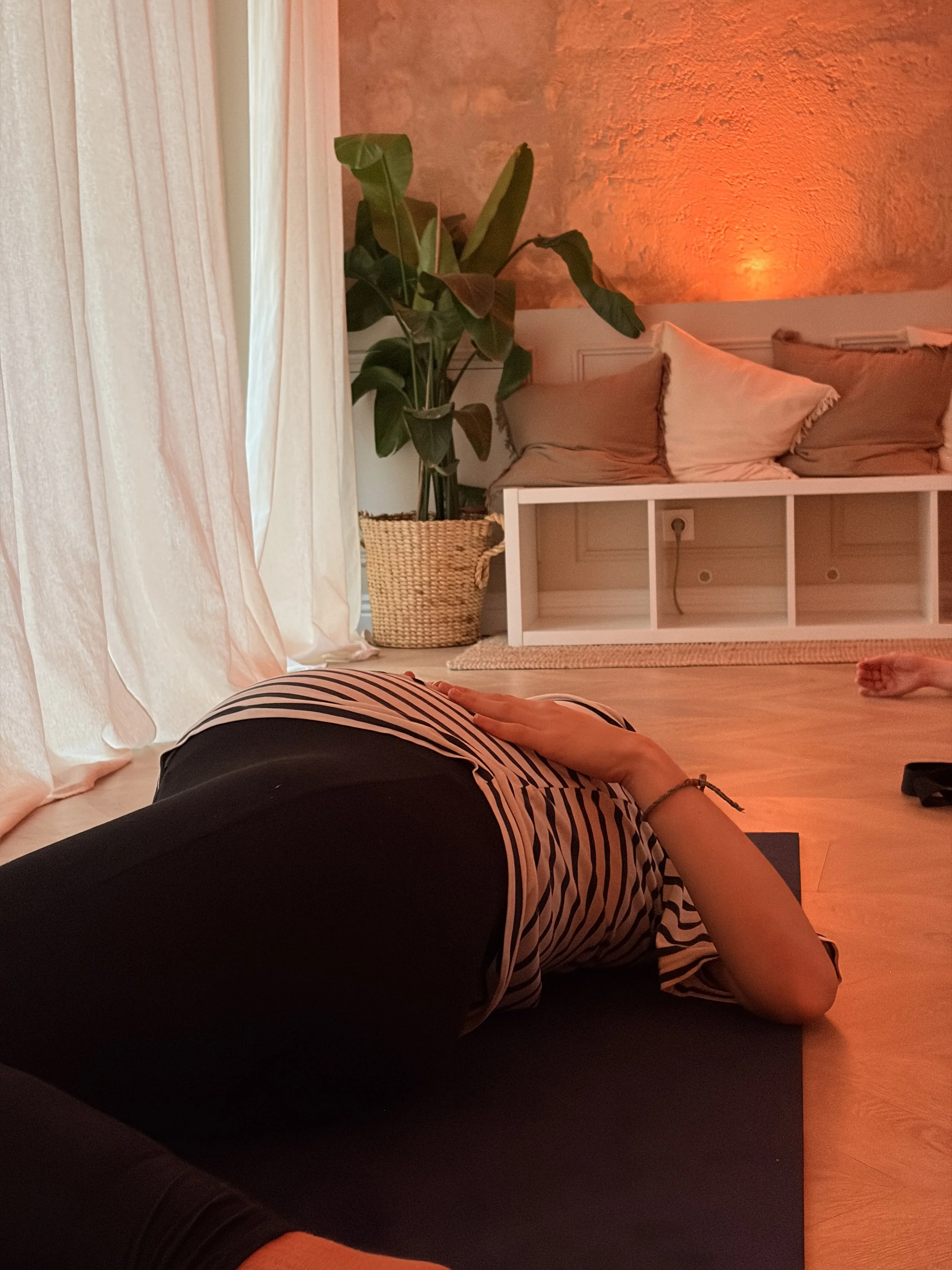 Une personne en position reclinée sur un tapis de yoga dans une pièce aux murs en pierre, avec un éclairage chaud. La personne porte une tenue à rayures et un bracelet, et l'angle de vue montre principalement le bras et la jambe.