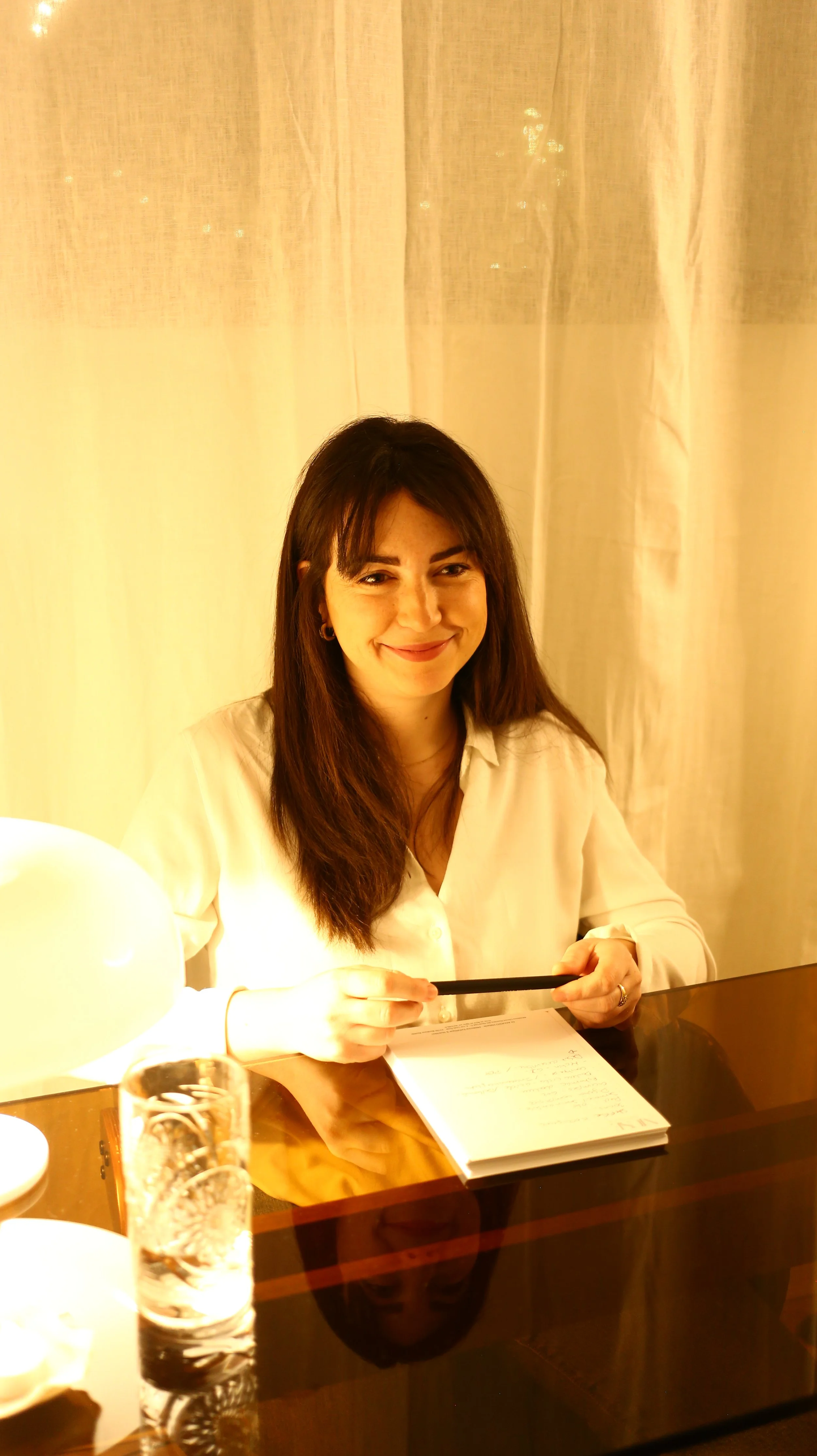 Femme souriante assise à une table, tenant un stylo, avec un carnet et un verre d'eau en premier plan, dans un environnement lumineux.