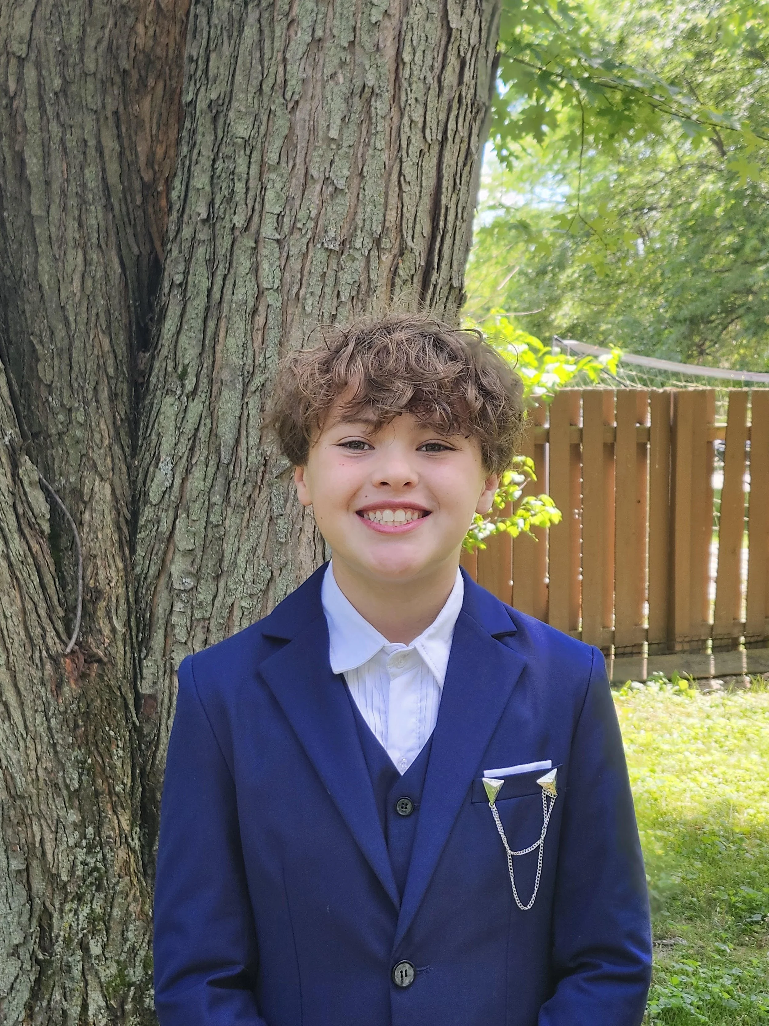 A young boy with curly hair smiling in front of a large tree with a textured bark, wearing a blue suit and white shirt, outdoors with green leaves and a wooden fence in the background.