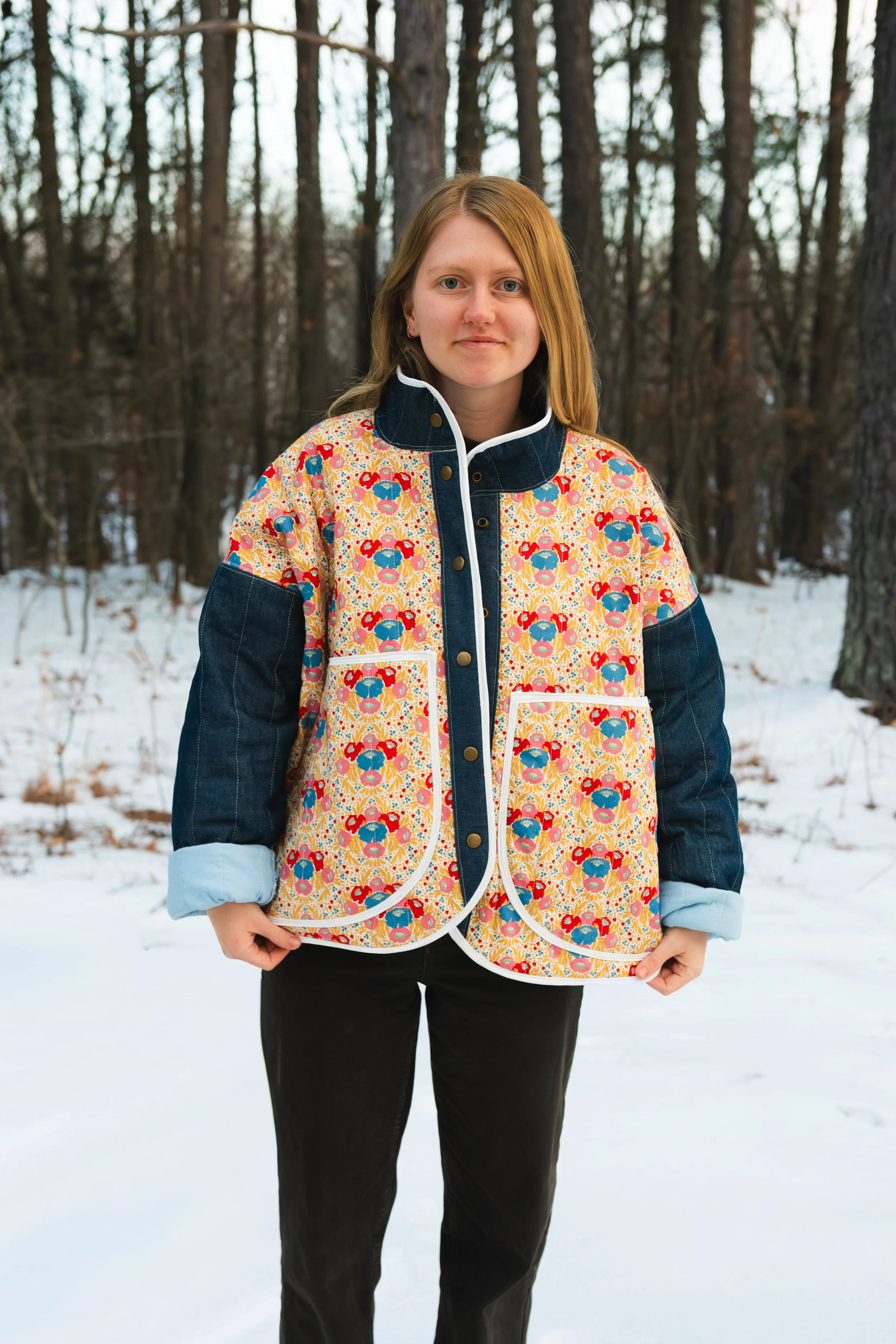 A young woman with red hair wearing a colorful floral jacket in a snowy forest.