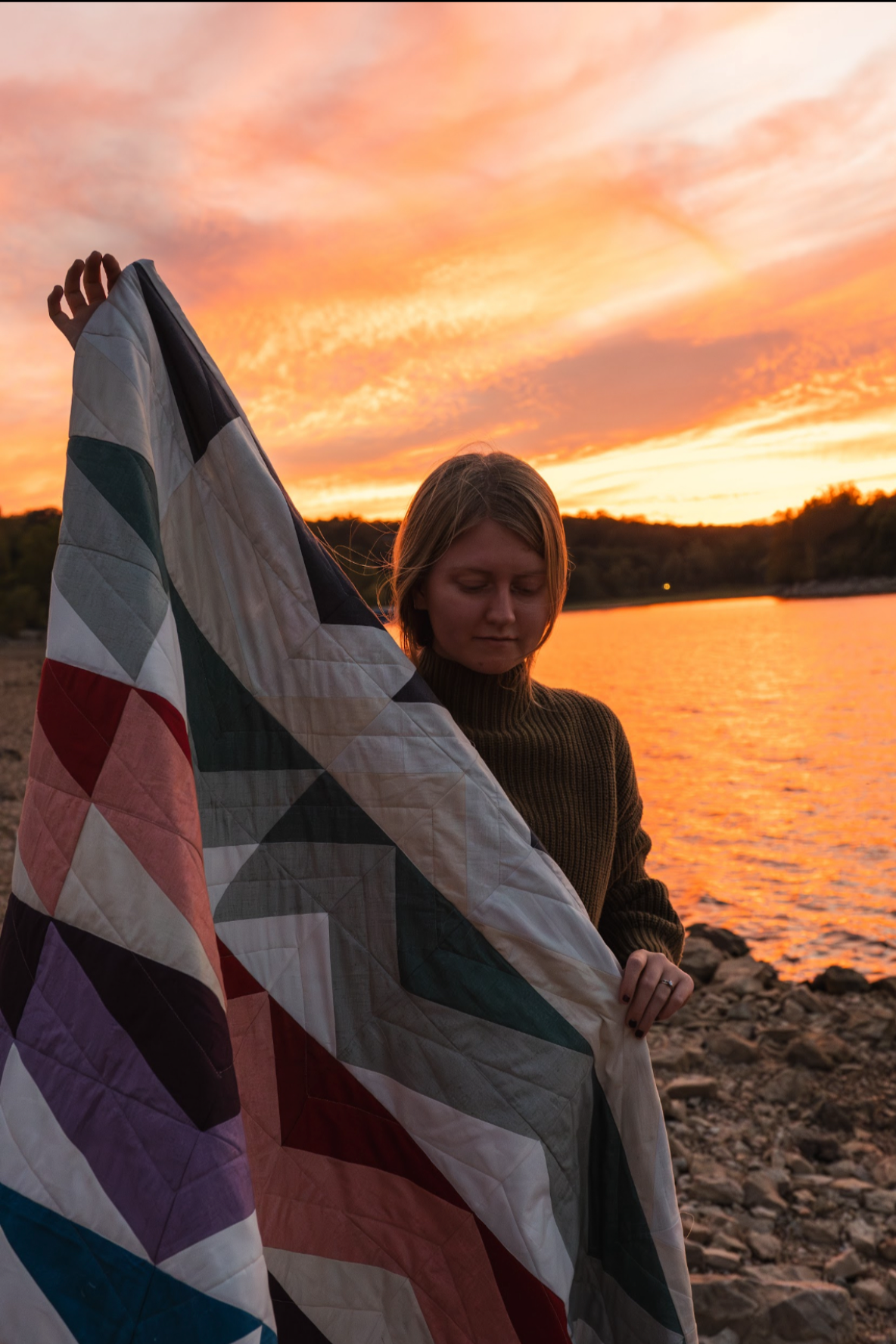 A woman holding a quilt or blanket with colorful geometric patterns, standing near a body of water during a vibrant sunset with orange and pink skies.