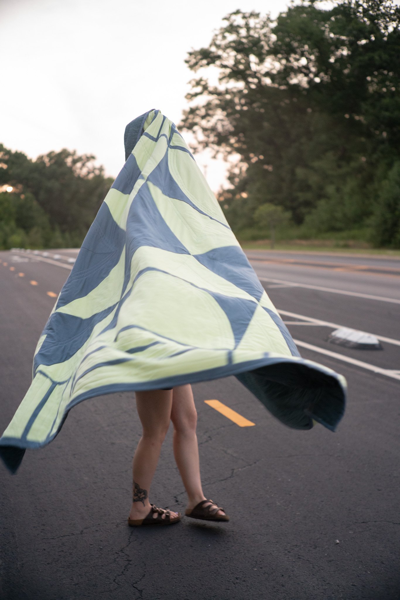 Person walking on a road with a large striped blanket or fabric draped over their head and shoulders, with trees visible in the background during dusk.