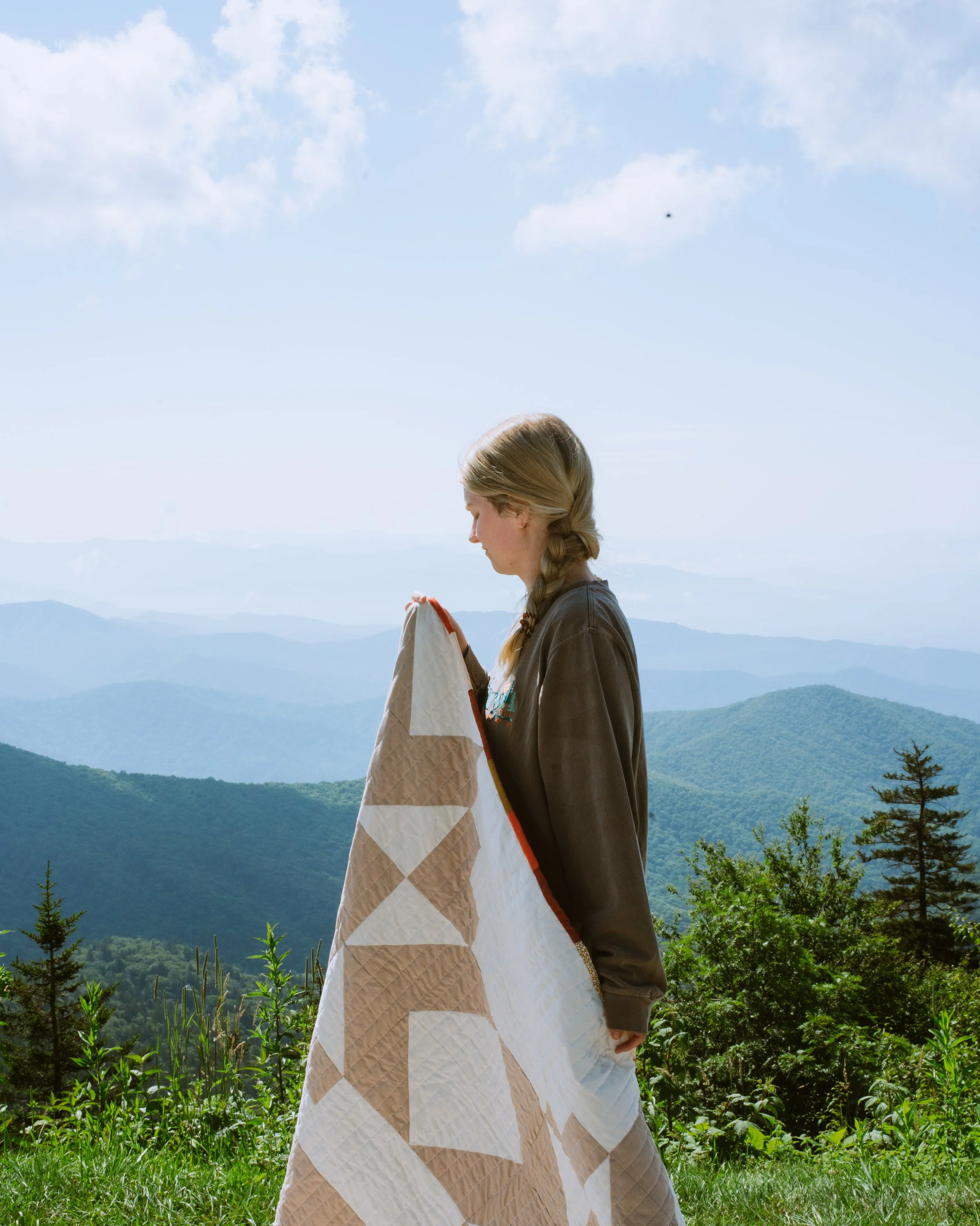 A young woman with blonde braided hair holding a bag, standing outdoors on a mountain with many layers of green trees and hills in the background, under a mostly cloudy sky.